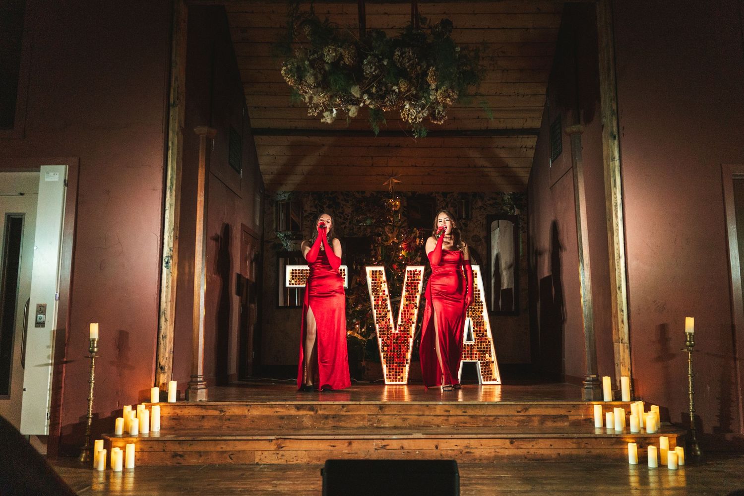 Concert performers in red dresses stand on stage with dramatic lighting and candles creating an atmospheric ambiance.