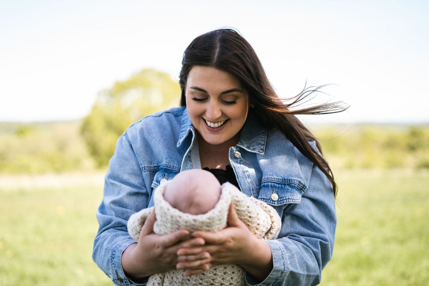 A newborn baby wrapped in a cream-colored knit blanket rests peacefully outdoors in the sunlight.