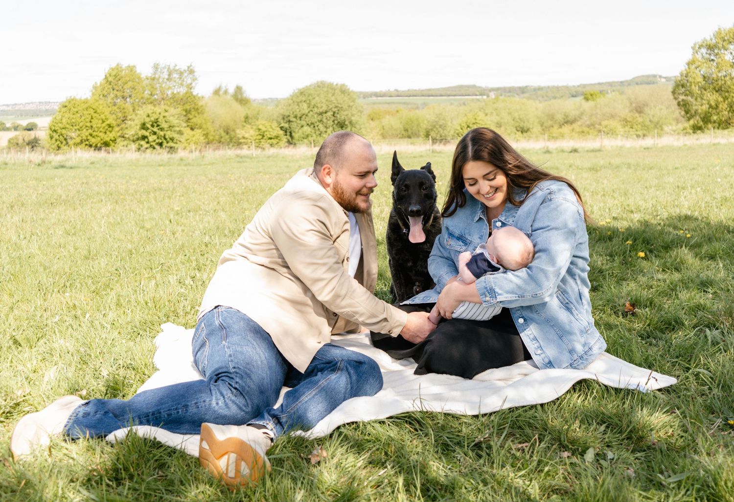 Family and pet dog lounging on white blanket in sunny meadow.