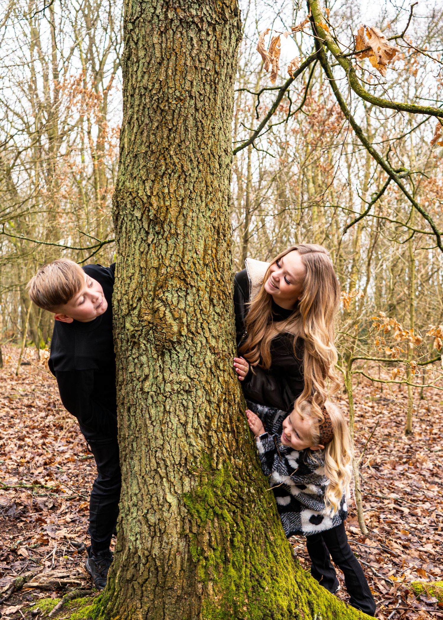 Candid forest photoshoot with people playing peek-a-boo around a mossy tree trunk.