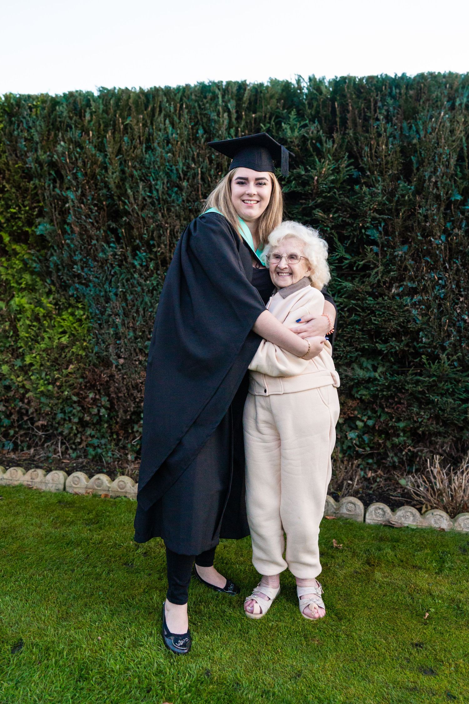 Graduate in academic dress and elderly relative pose together on garden lawn during celebration.