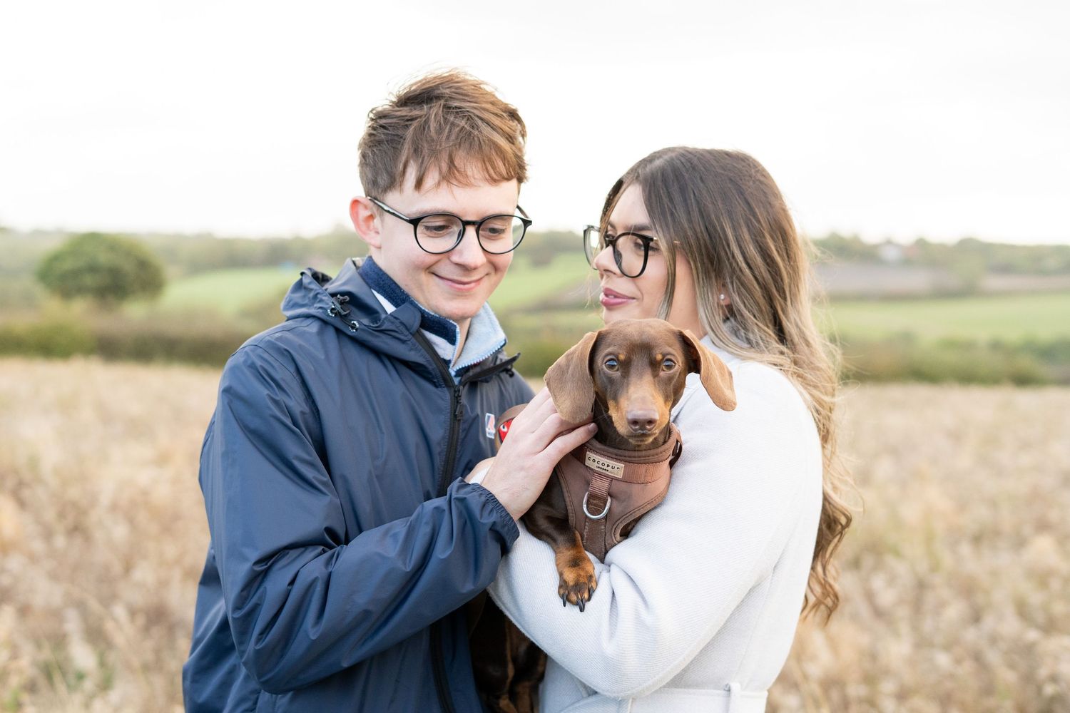 A couple snuggling with their brown dachshund puppy in an autumn field.