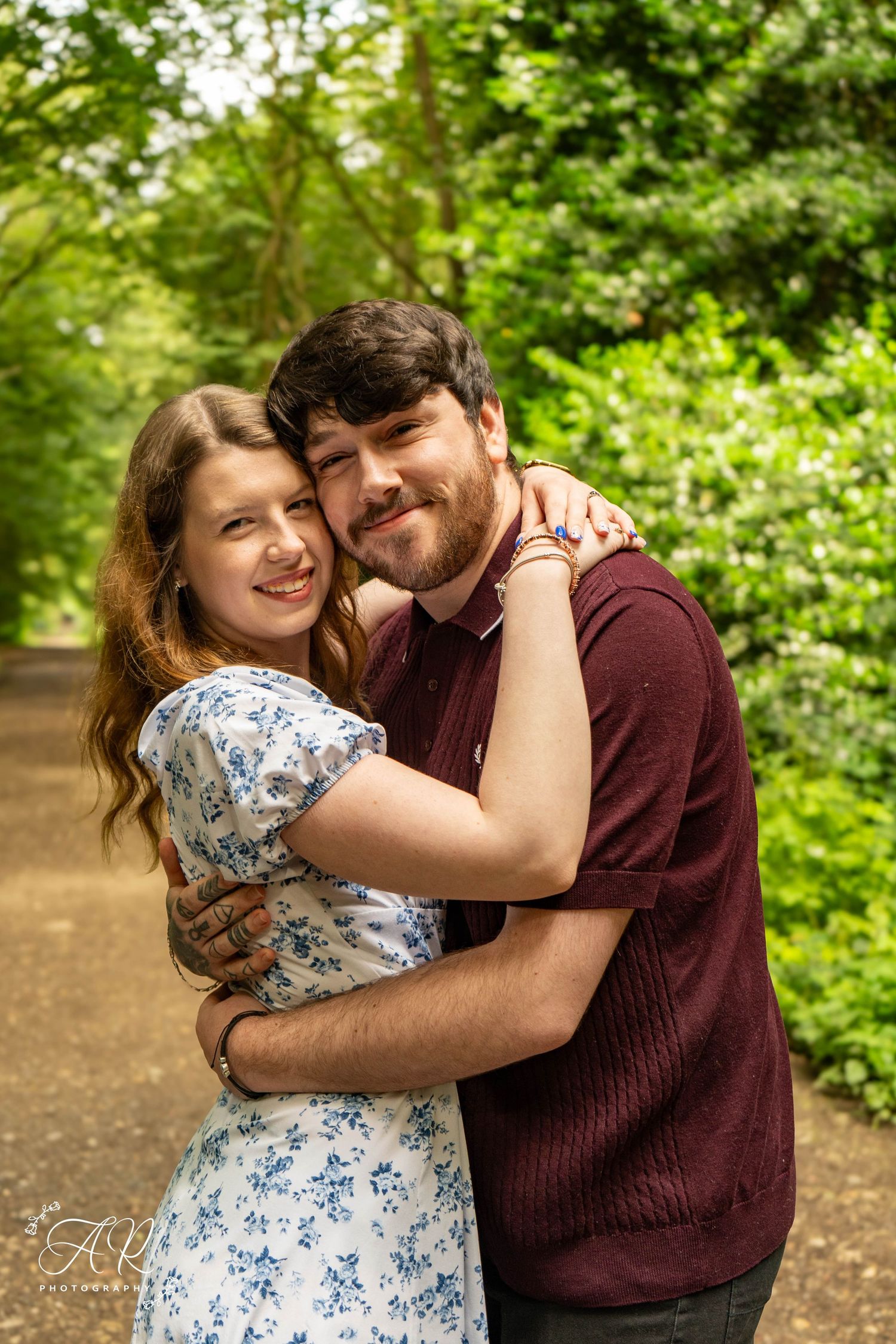 A couple embraces lovingly in a lush green garden setting wearing burgundy and floral print outfits.
