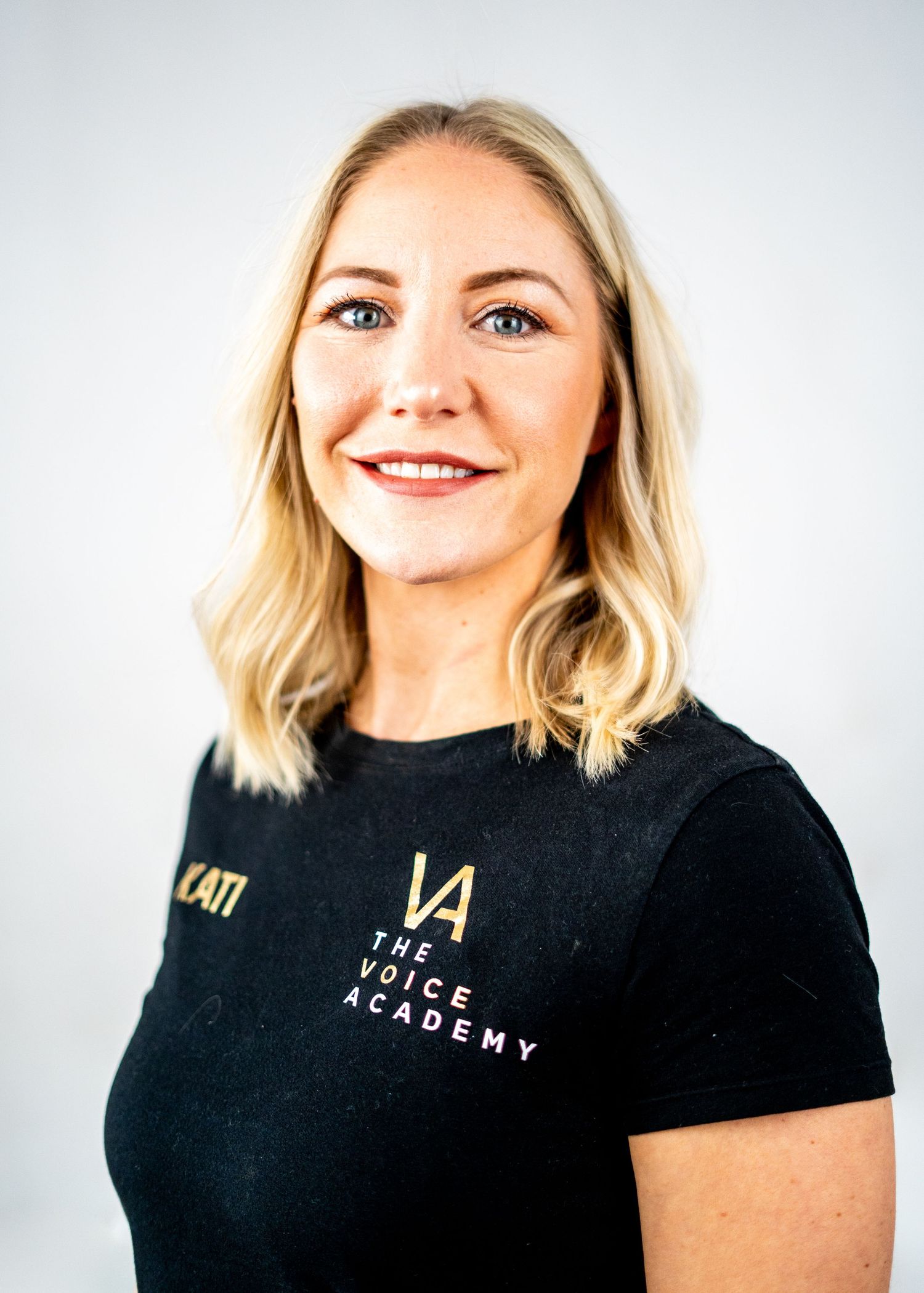 Professional headshot of a person in black Voice Academy shirt with shoulder-length blonde hair.