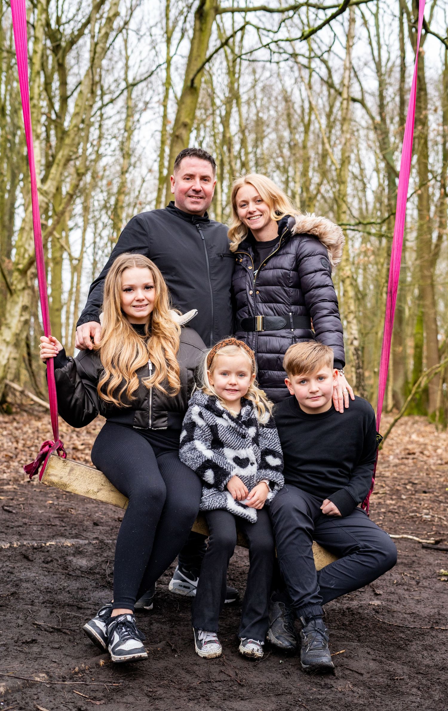 Family group sitting together on forest ground with pink ribbons visible in background.