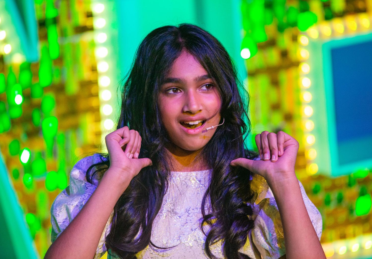 Young performer in white lace dress gestures expressively against vibrant green and yellow stage lighting.
