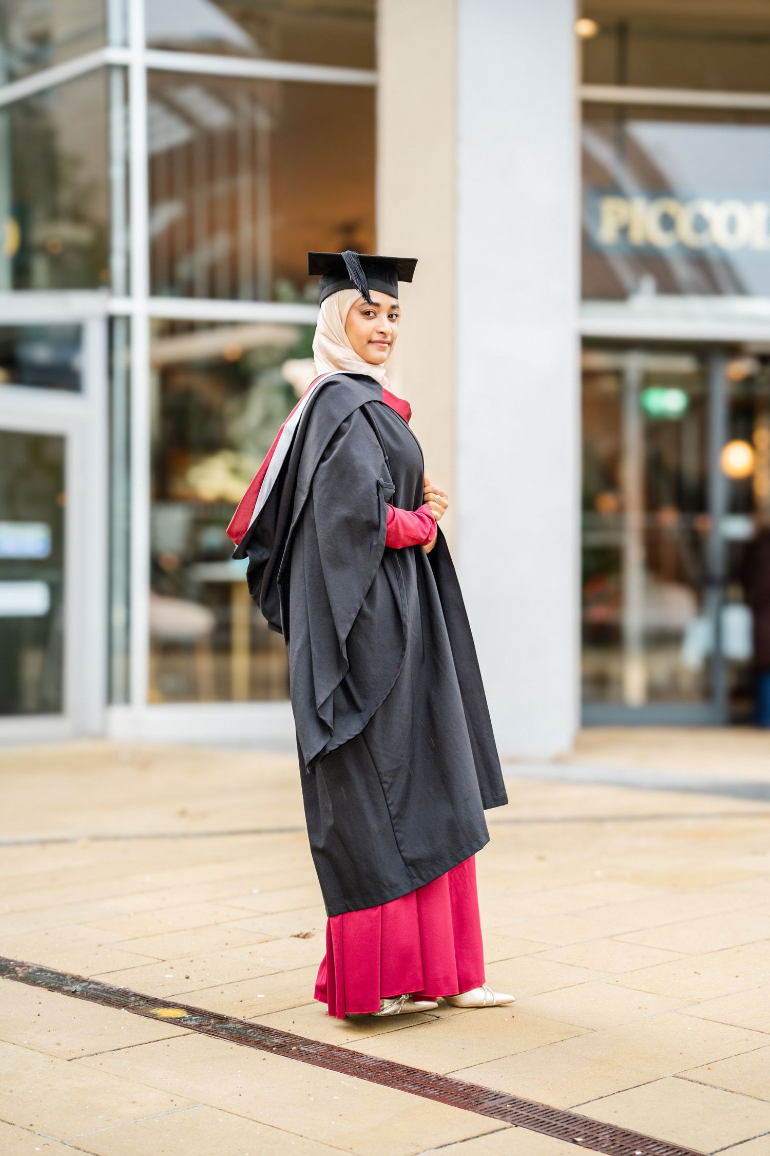 Graduate in academic dress stands in front of campus building during photo session.