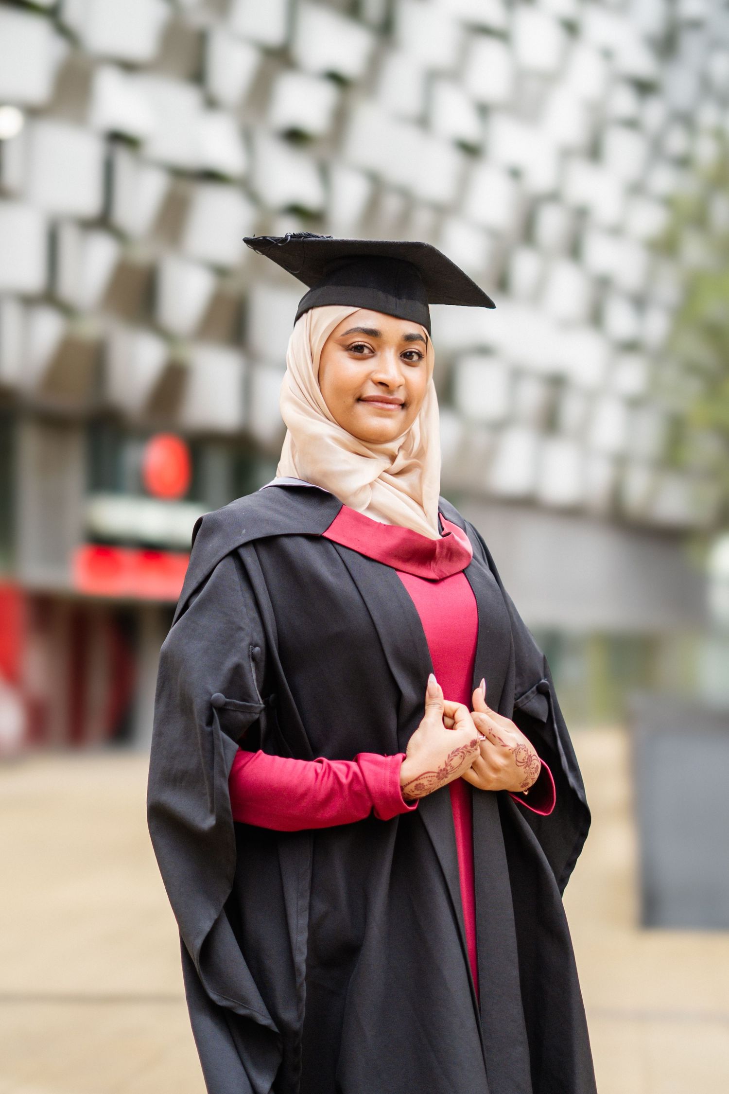 Graduate wearing black academic regalia and red dress poses against modern architectural background.