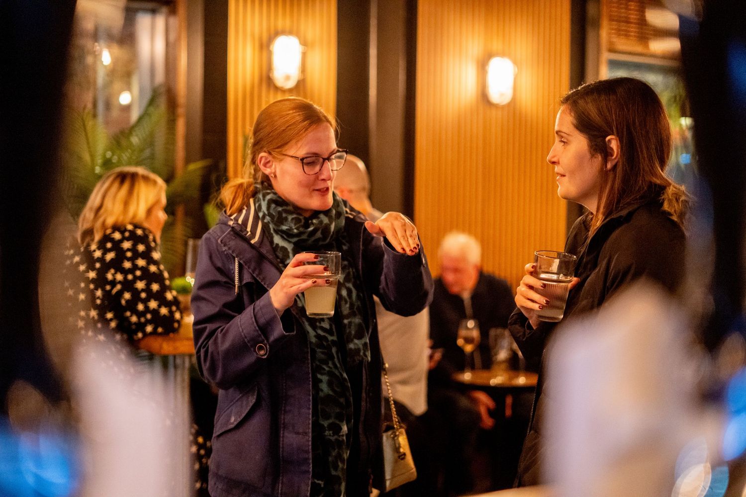 Patrons engaged in conversation at a dimly lit bar with wall sconces creating intimate atmosphere.