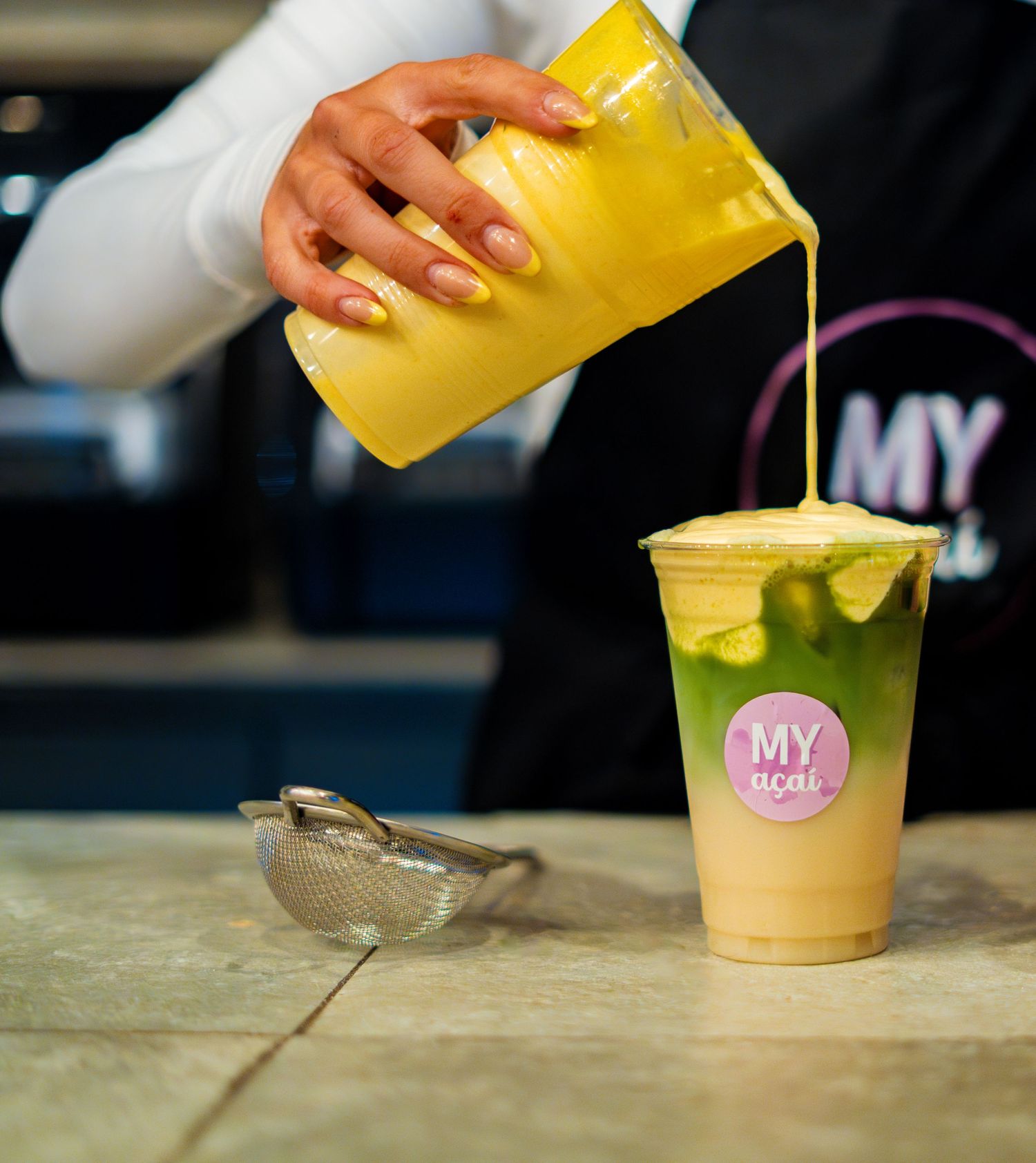 Yellow liquid being poured into a pink branded cup containing a creamy beverage.