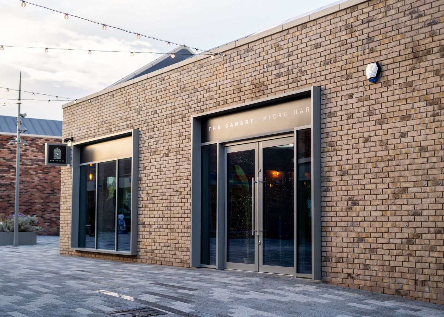 Modern brick building exterior with glass doors and string lights overhead.