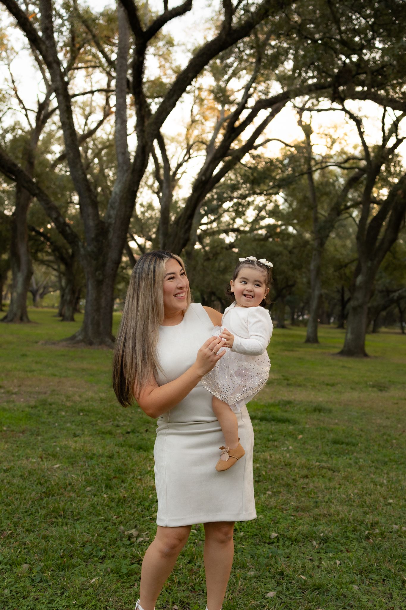 A joyful moment shared between parent and child in white dresses among oak trees.