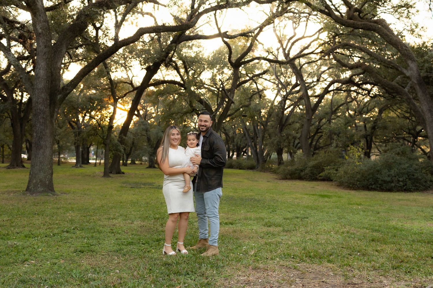 A couple poses together on grass beneath towering oak trees at sunset.