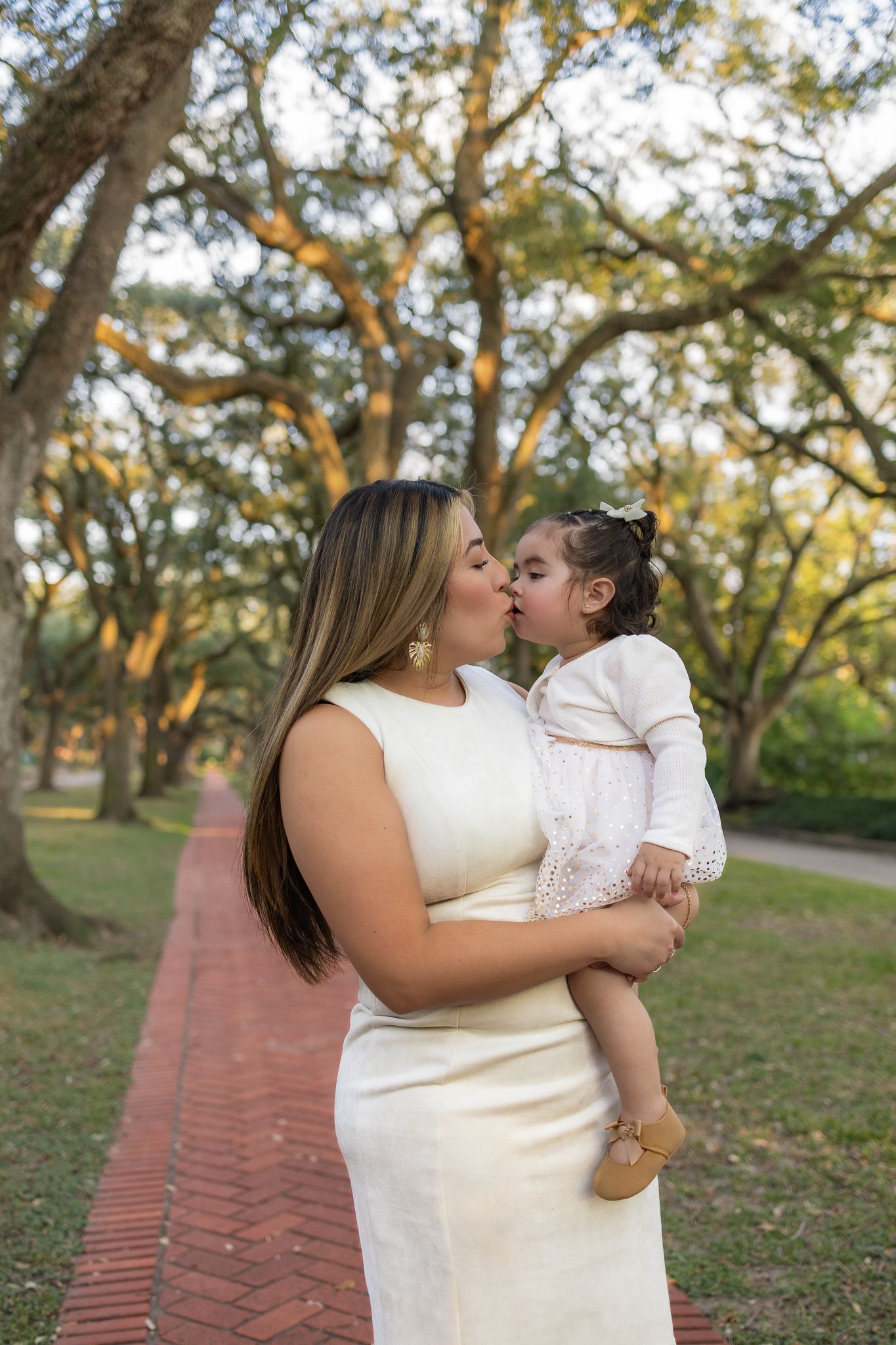 A tender moment captured between parent and child in a white dress against a backdrop of sunlit trees.