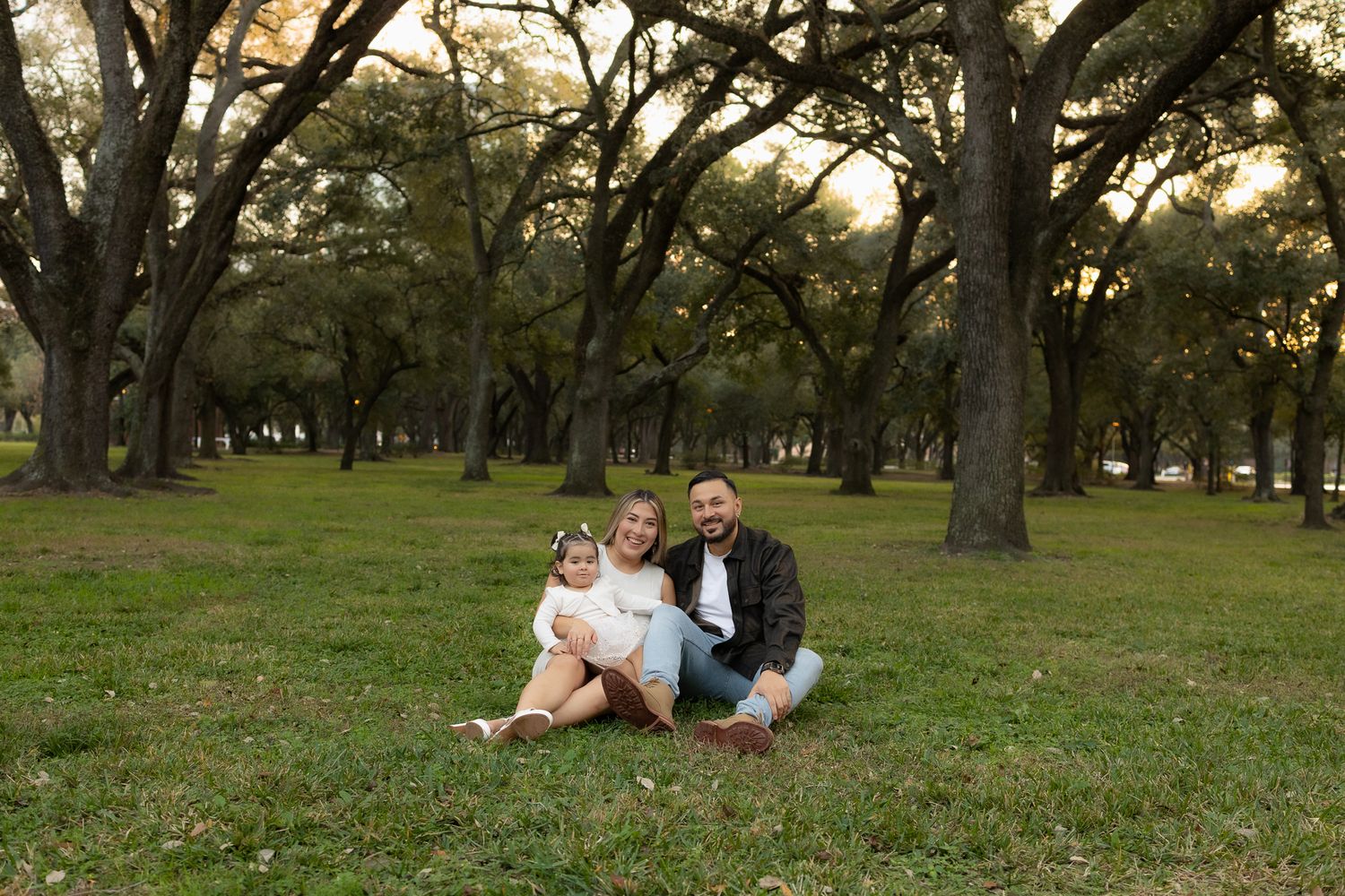 A family relaxes together on grass beneath a canopy of oak trees.