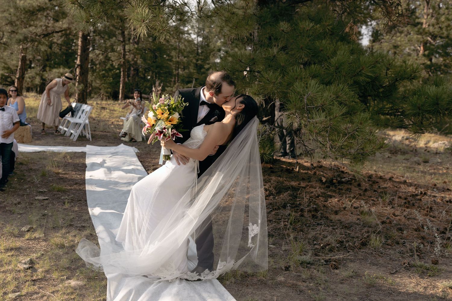 Bride and groom kissing during their outdoor wedding ceremony surrounded by trees and guests
