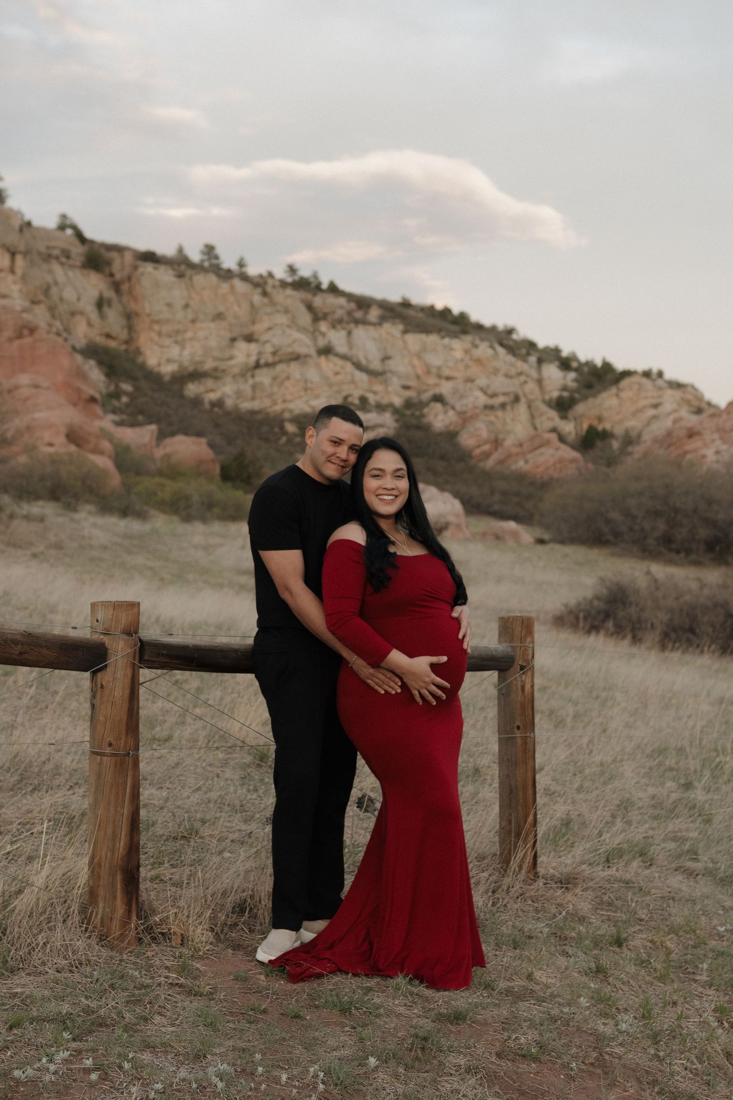 Expecting couple embracing outdoors in a red maternity dress, surrounded by mountains scenery