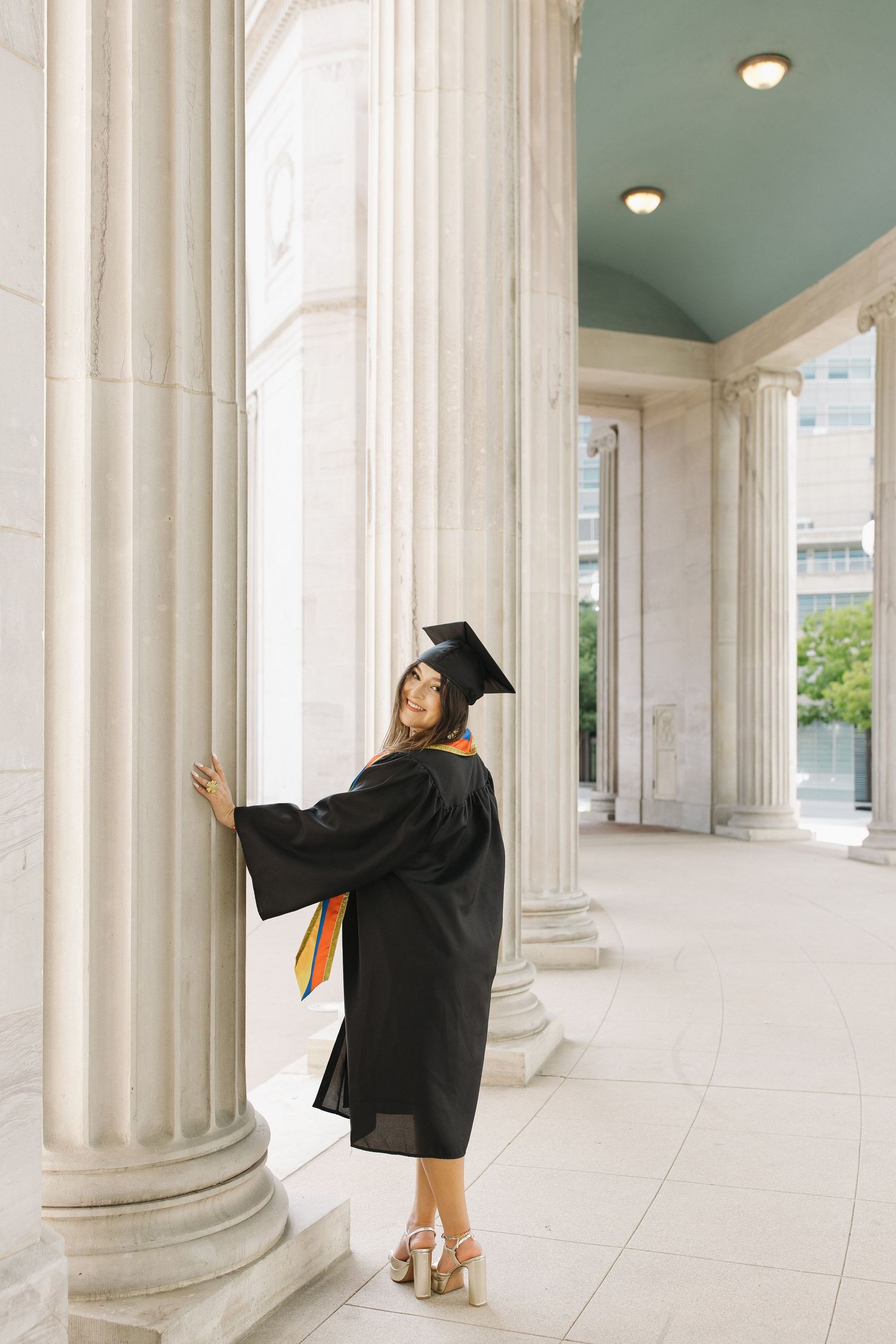 Graduation portrait of a young woman in a cap and gown posing beside elegant columns