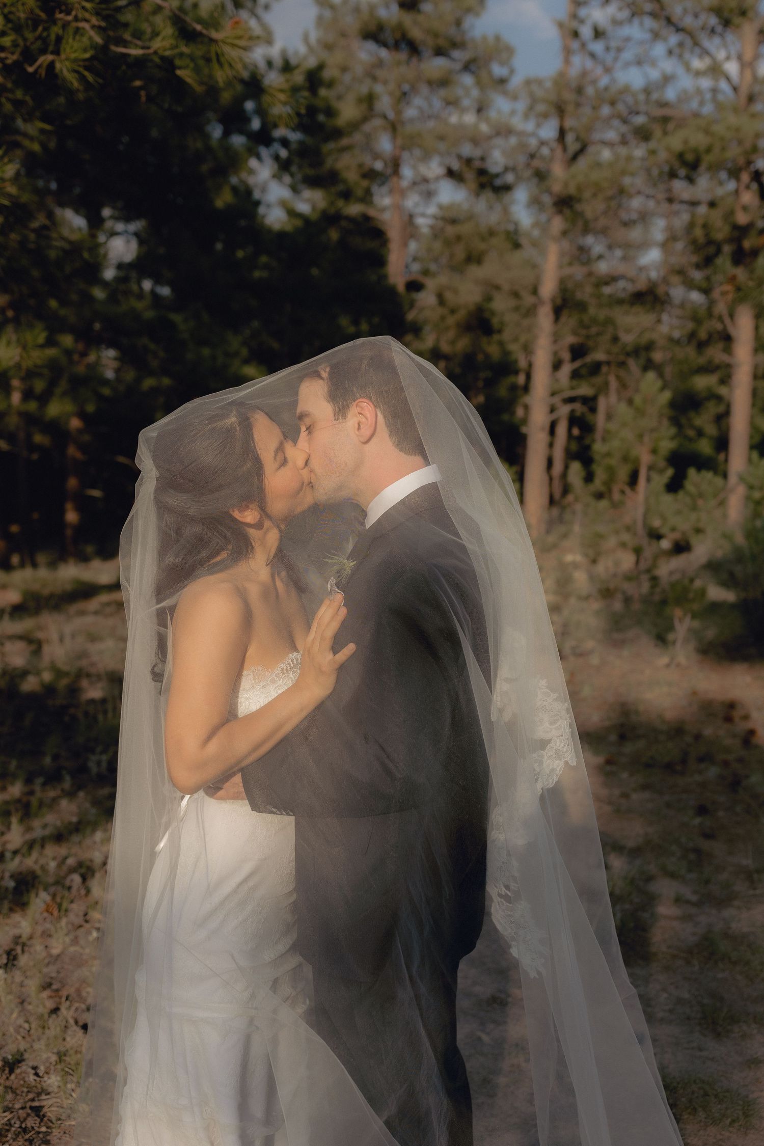 Bride and groom kissing under the bride’s veil in a wooded setting