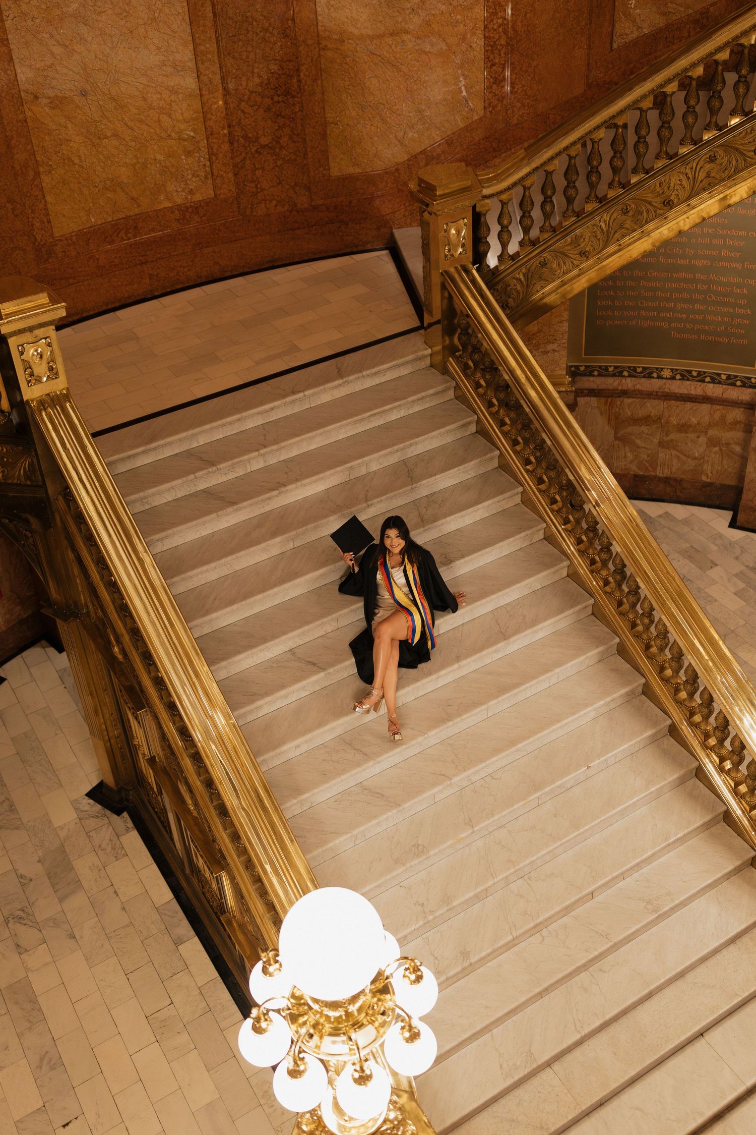 Graduation portrait of a young woman in a cap and gown standing on an elegant grand staircase, photographed from above