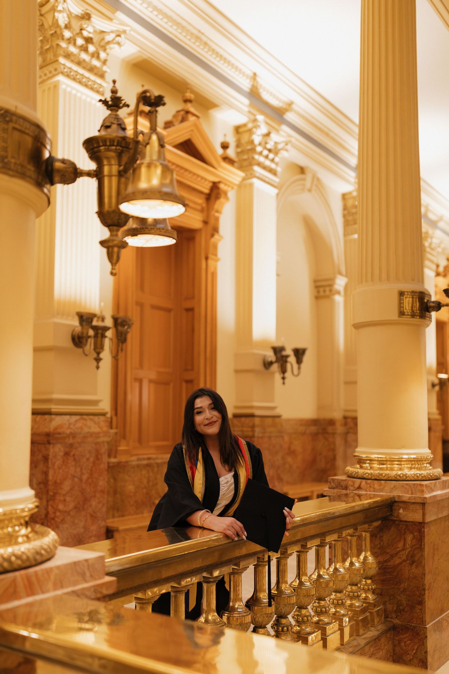 Young woman in graduation attire sitting beside an ornate staircase during an elegant graduation portrait session