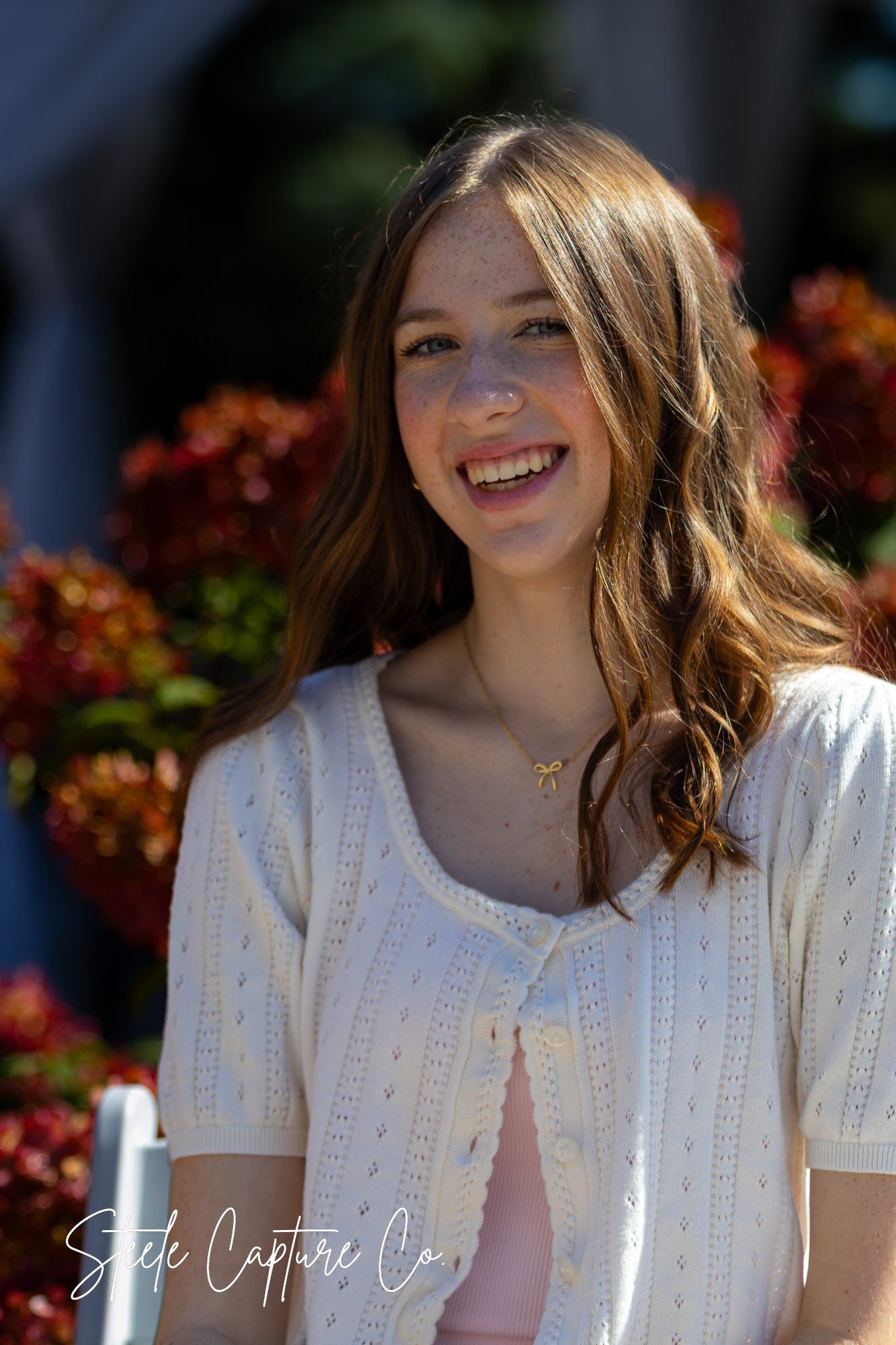 Senior girl in cream cardigan smiling in front of red flowers