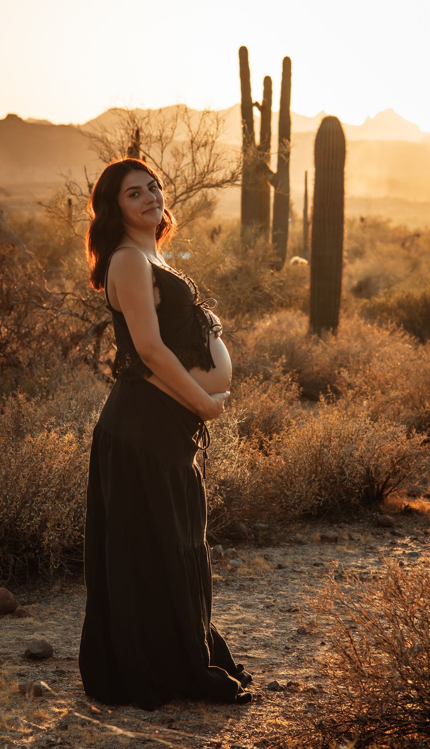 Pregnant woman in black dress holding baby bump at golden hour in Arizona desert with saguaro cacti backdrop.