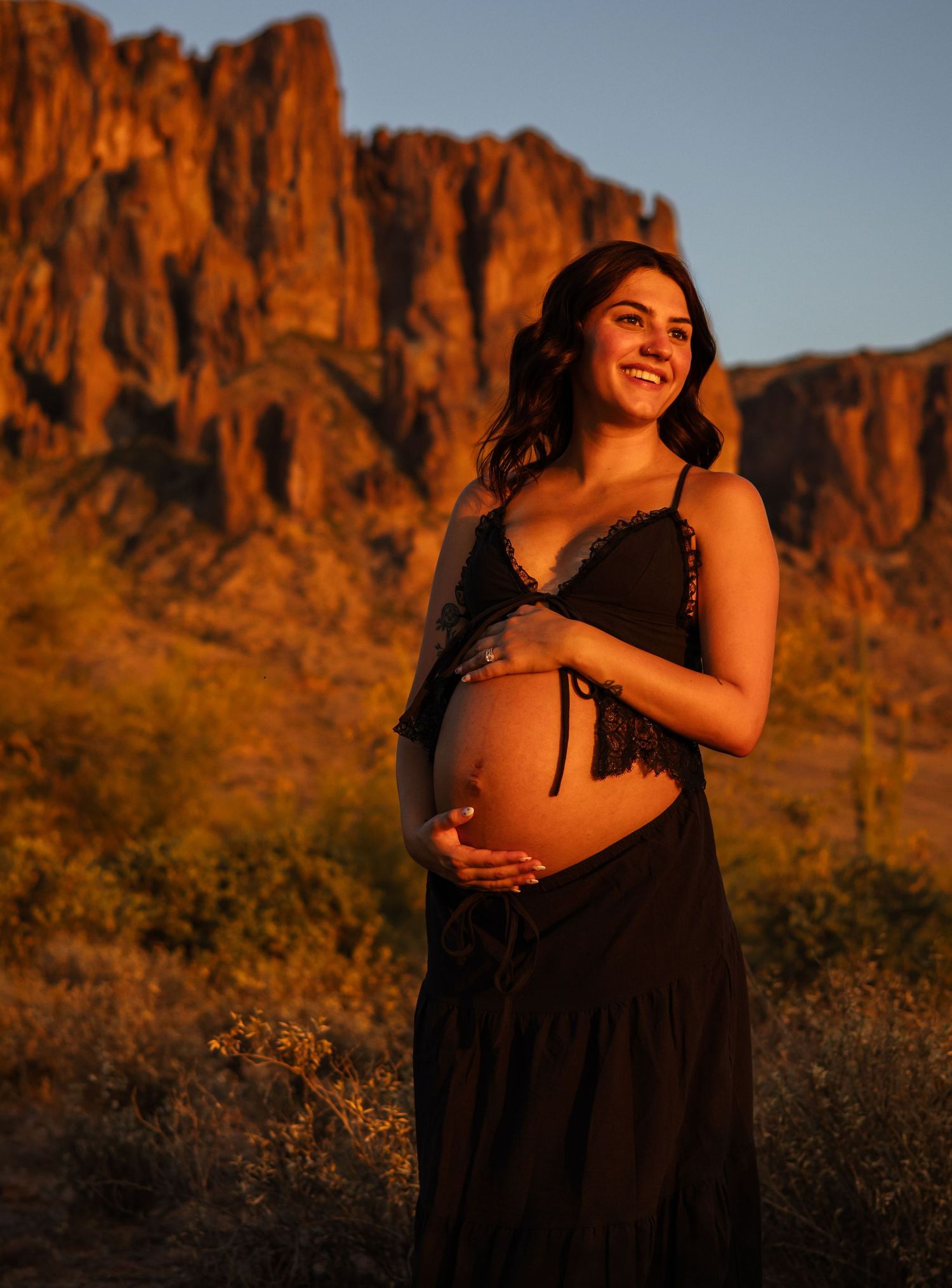 Smiling pregnant woman in black dress cradling baby bump at golden hour with rocky desert mountains in background.