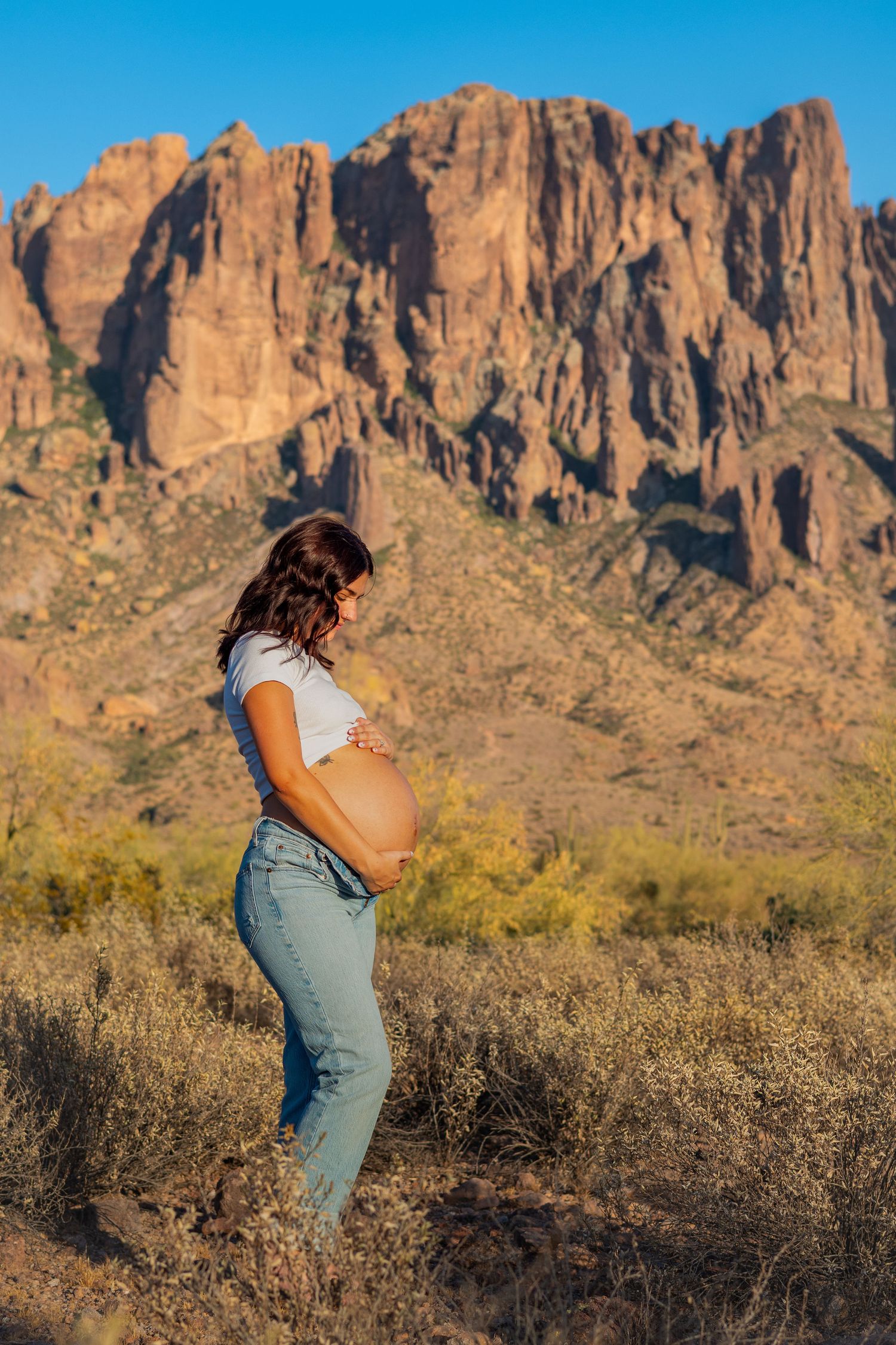 Pregnant woman in jeans cradling her baby bump outdoors with dramatic desert mountain landscape in background.