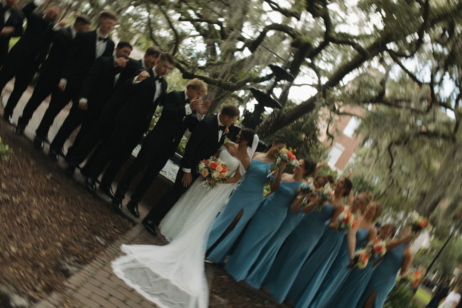 Wedding party poses outdoors under oak trees, groomsmen in black tuxedos and bridesmaids in teal dresses flanking the bride and groom.