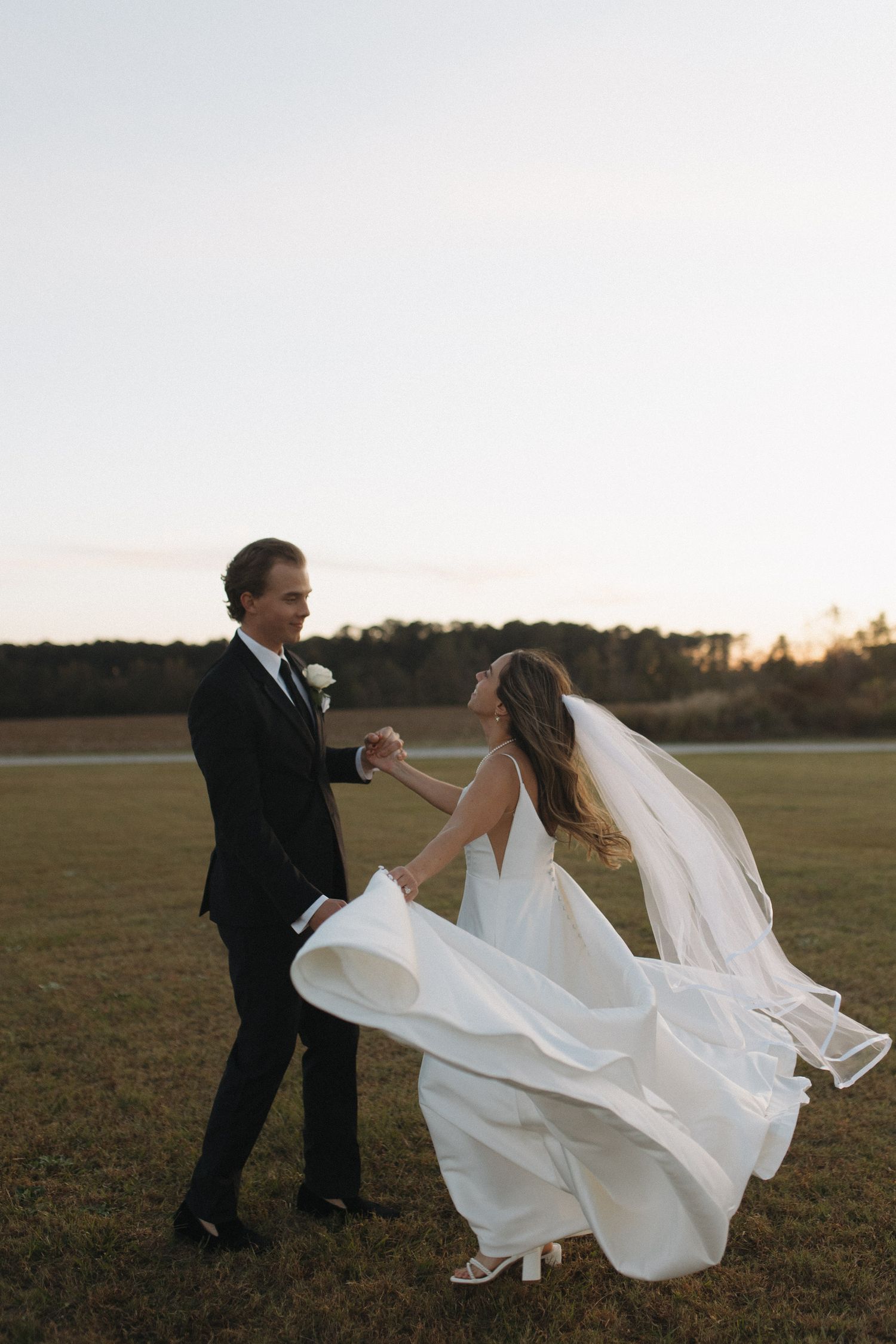 Bride and groom dancing in a field at sunset, her white gown and veil flowing in the wind.