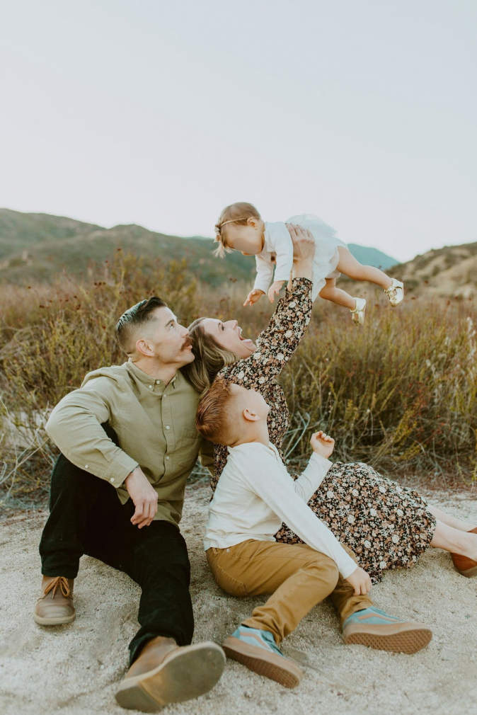 A joyful family of four plays outdoors, mom lifting baby in the air while dad and son sit on sandy ground with hills behind.