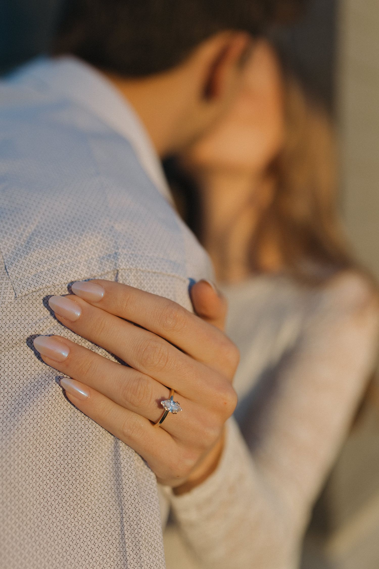 Close-up of a womans hand with a diamond engagement ring resting on her partners shoulder.