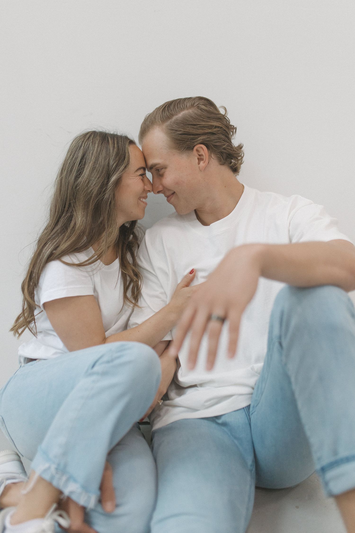 Couple in matching white tees and jeans sitting on floor, touching foreheads and smiling at each other.