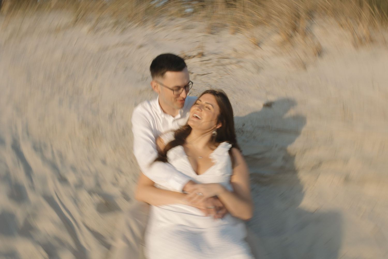 Happy couple embraces and laughs together during romantic beach engagement photo session at sunset.