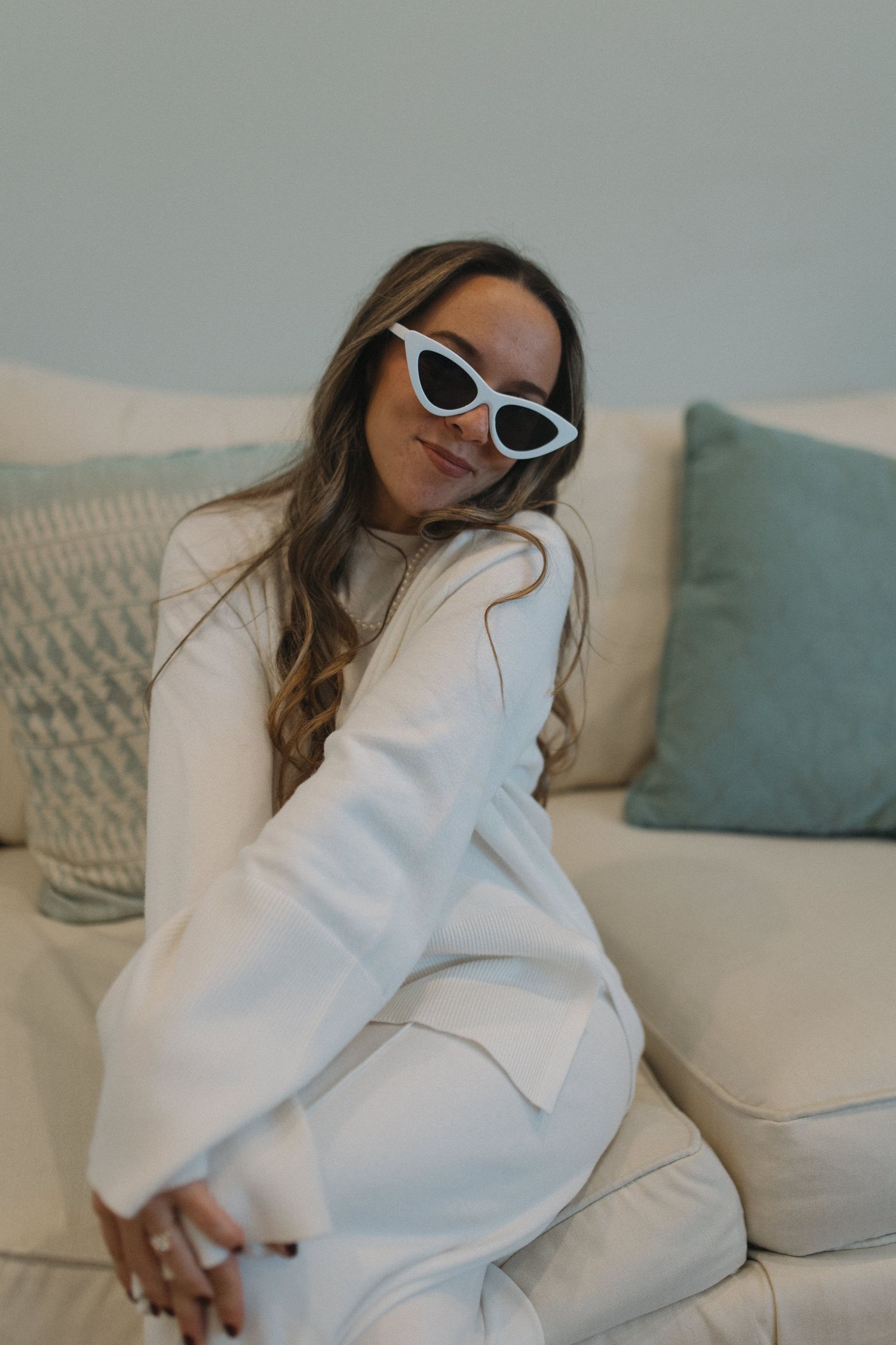 Stylish woman in a white outfit wearing cat-eye sunglasses relaxing on a cream sofa.