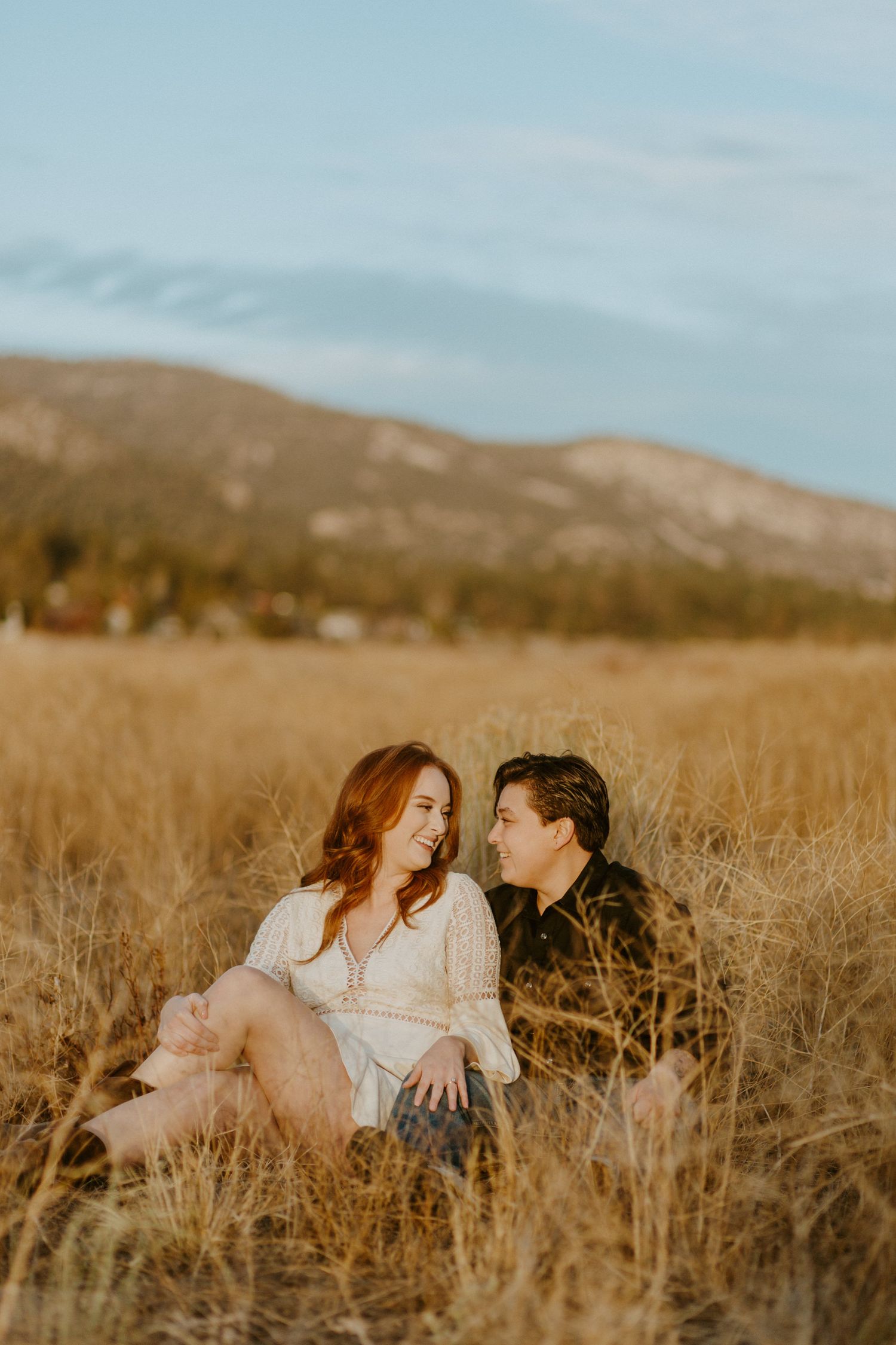 Couple sitting in golden dry grass field gazing at each other with mountain backdrop.