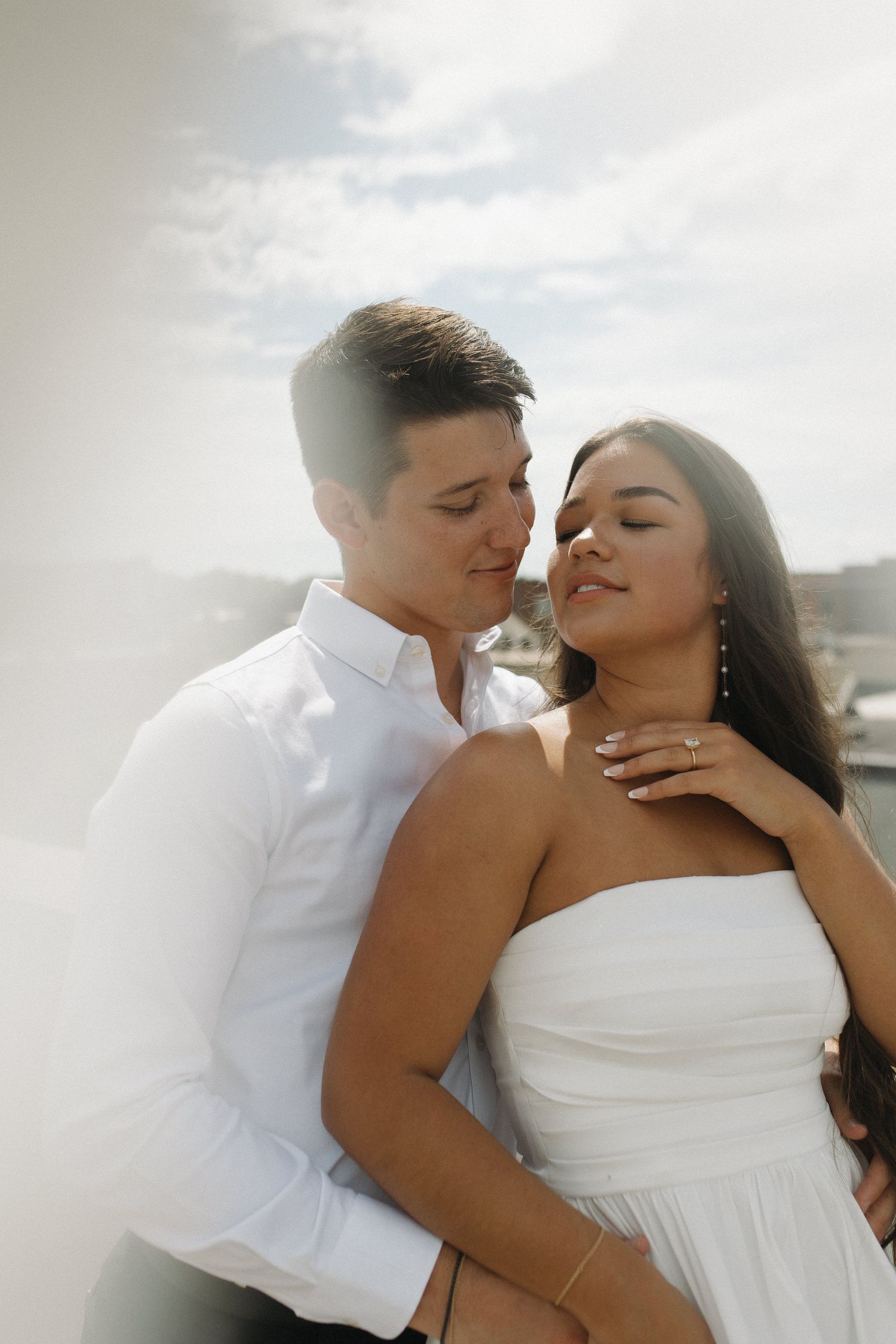 Romantic couple in white outfits embracing tenderly against a bright cloudy sky backdrop.