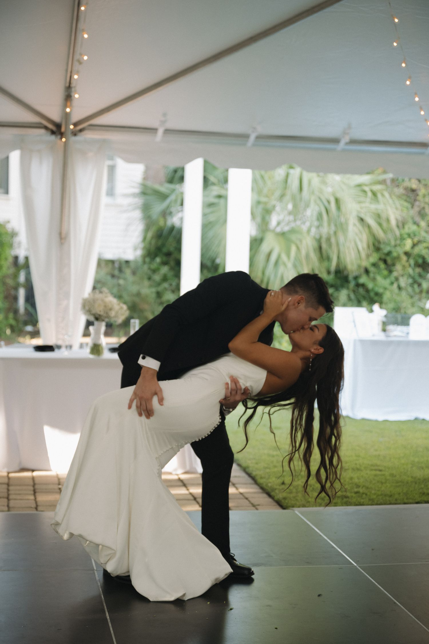 Groom dips bride for a kiss during their first dance under a white tent at outdoor reception.