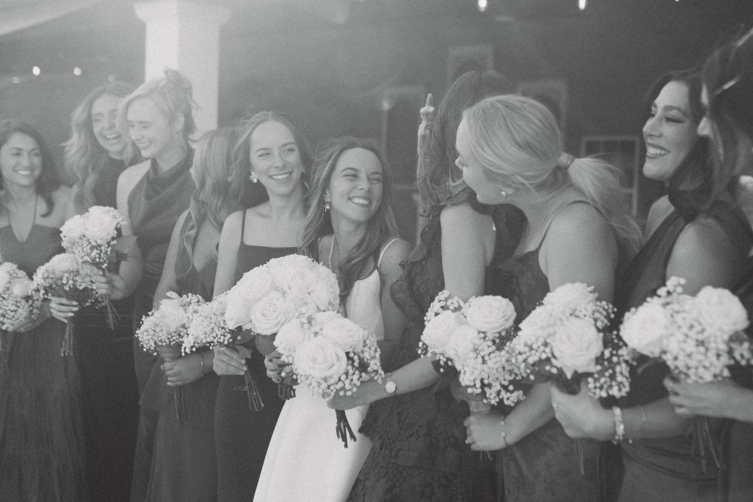 Black and white photo of a bride and bridesmaids laughing together holding white bouquets at a wedding celebration.