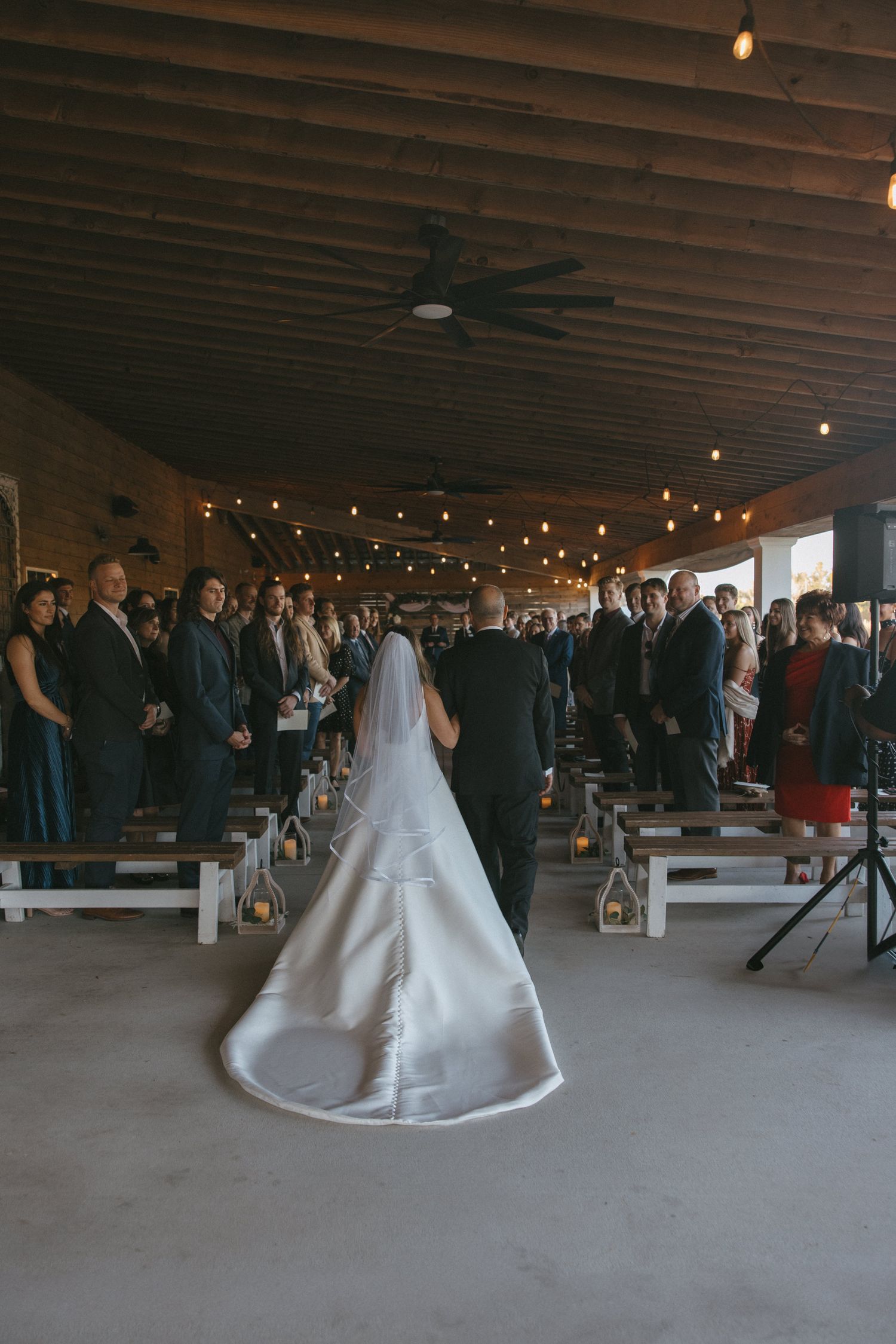 Bride walking down the aisle in a white gown and veil at a rustic indoor wedding venue with string lights.