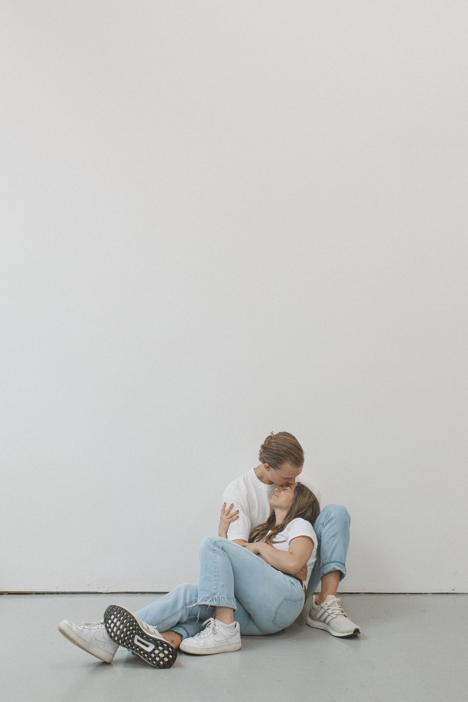 Couple in white tees and jeans sitting on floor, embracing tenderly against a white wall.