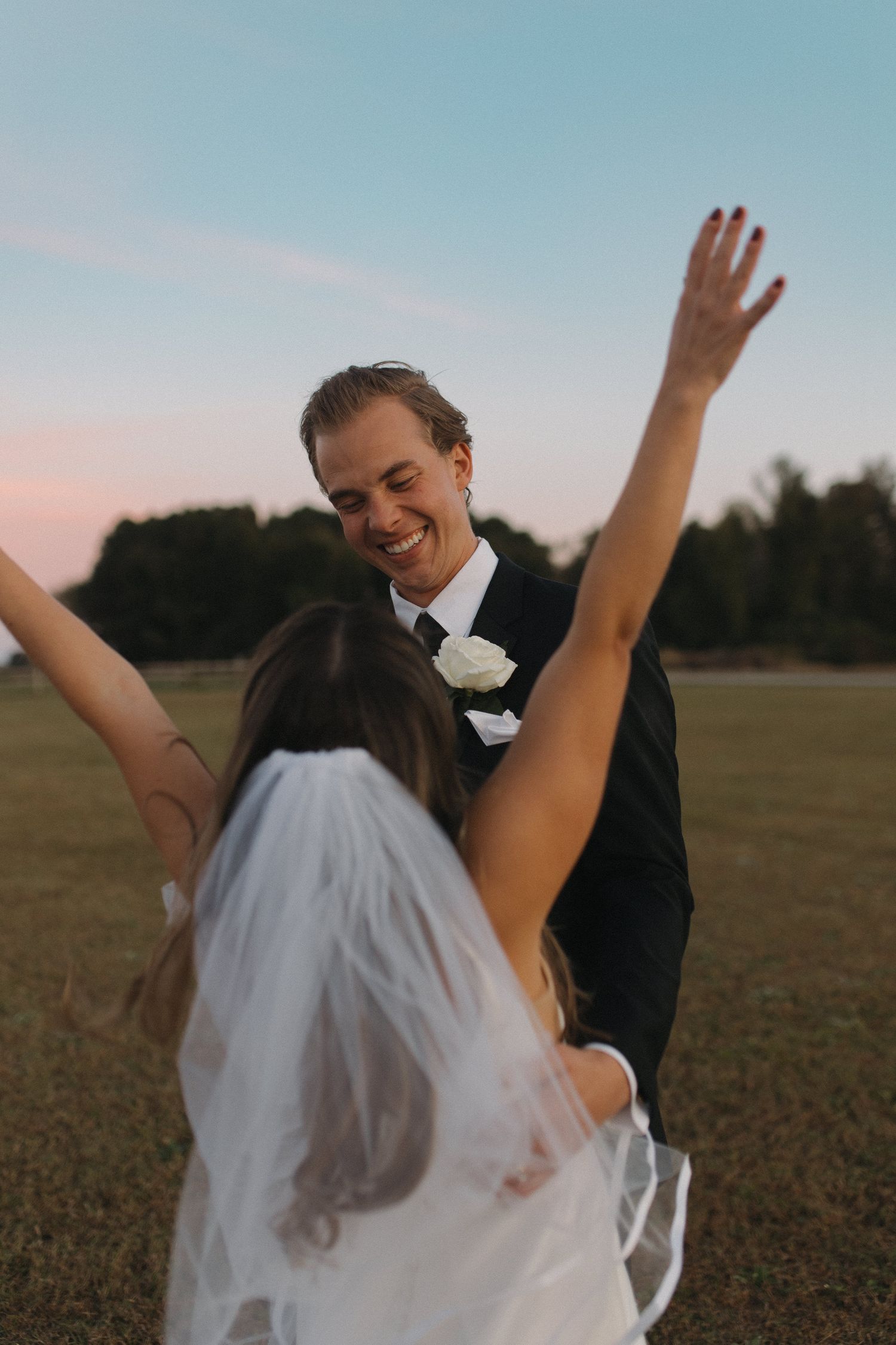Joyful newlyweds celebrate outdoors at sunset, groom in black suit lifting bride in white dress and veil.