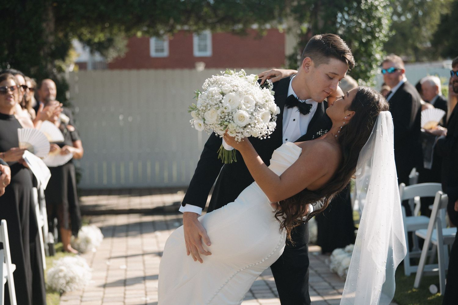 Groom dips bride in white gown and veil for a romantic kiss at the end of their outdoor wedding ceremony aisle.