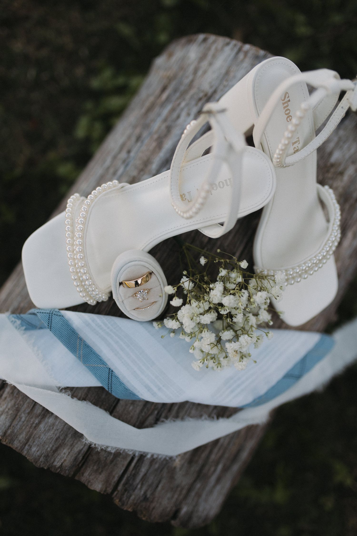 White pearl-adorned bridal heels with wedding rings and baby's breath on a rustic wooden surface with a blue ribbon.