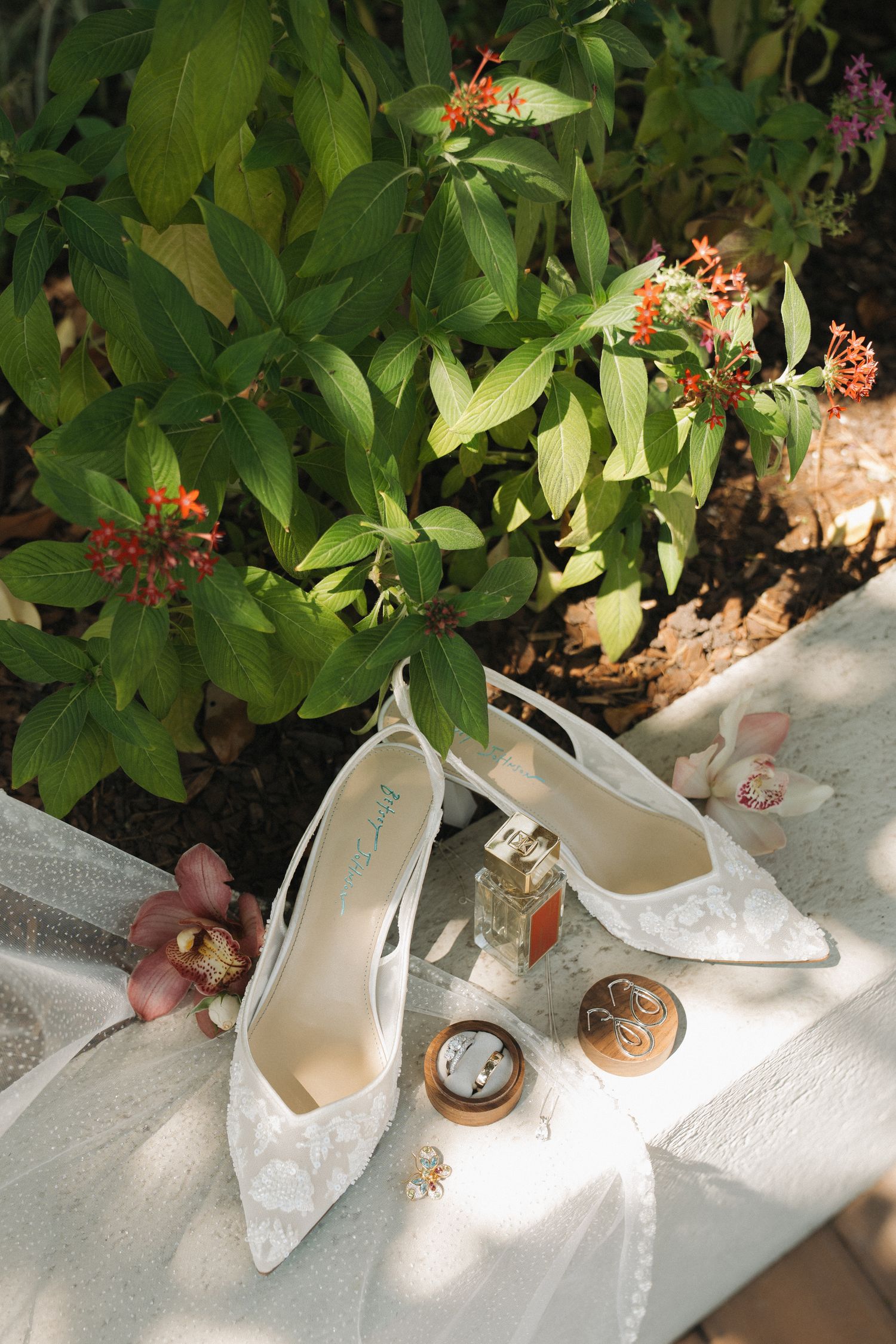 White lace bridal heels displayed with perfume and jewelry on fabric near tropical flowers.