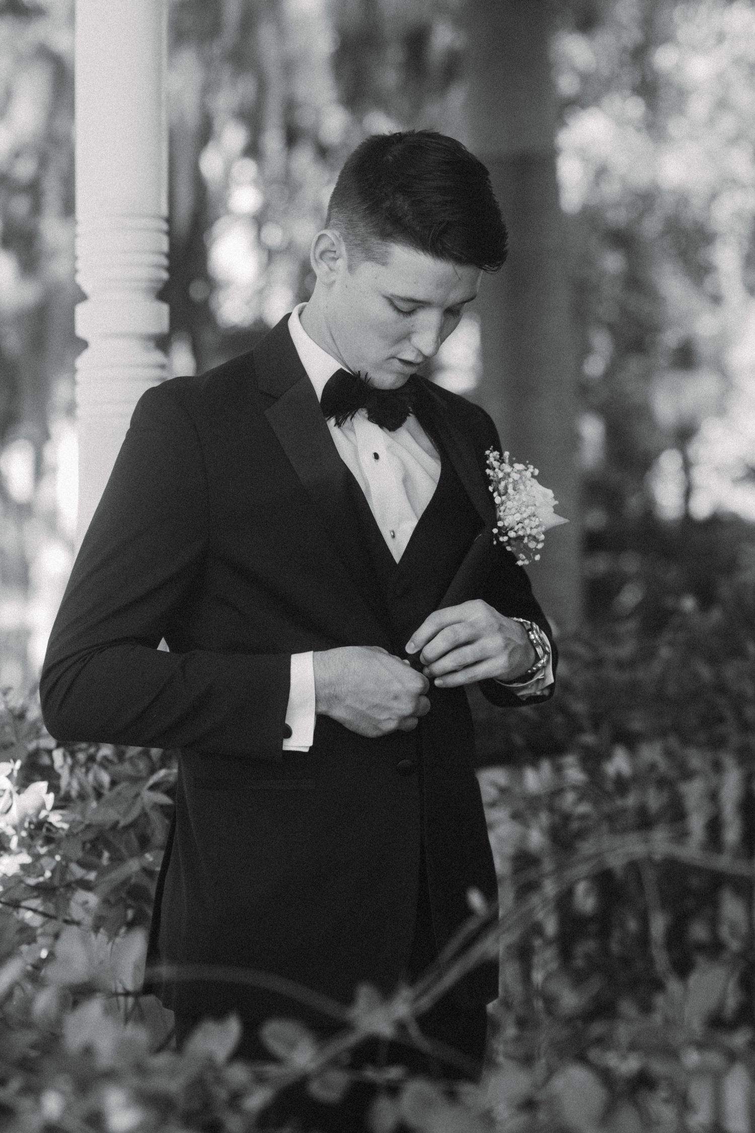 Groom in black tuxedo fastening jacket button with floral boutonniere outdoors in black and white.