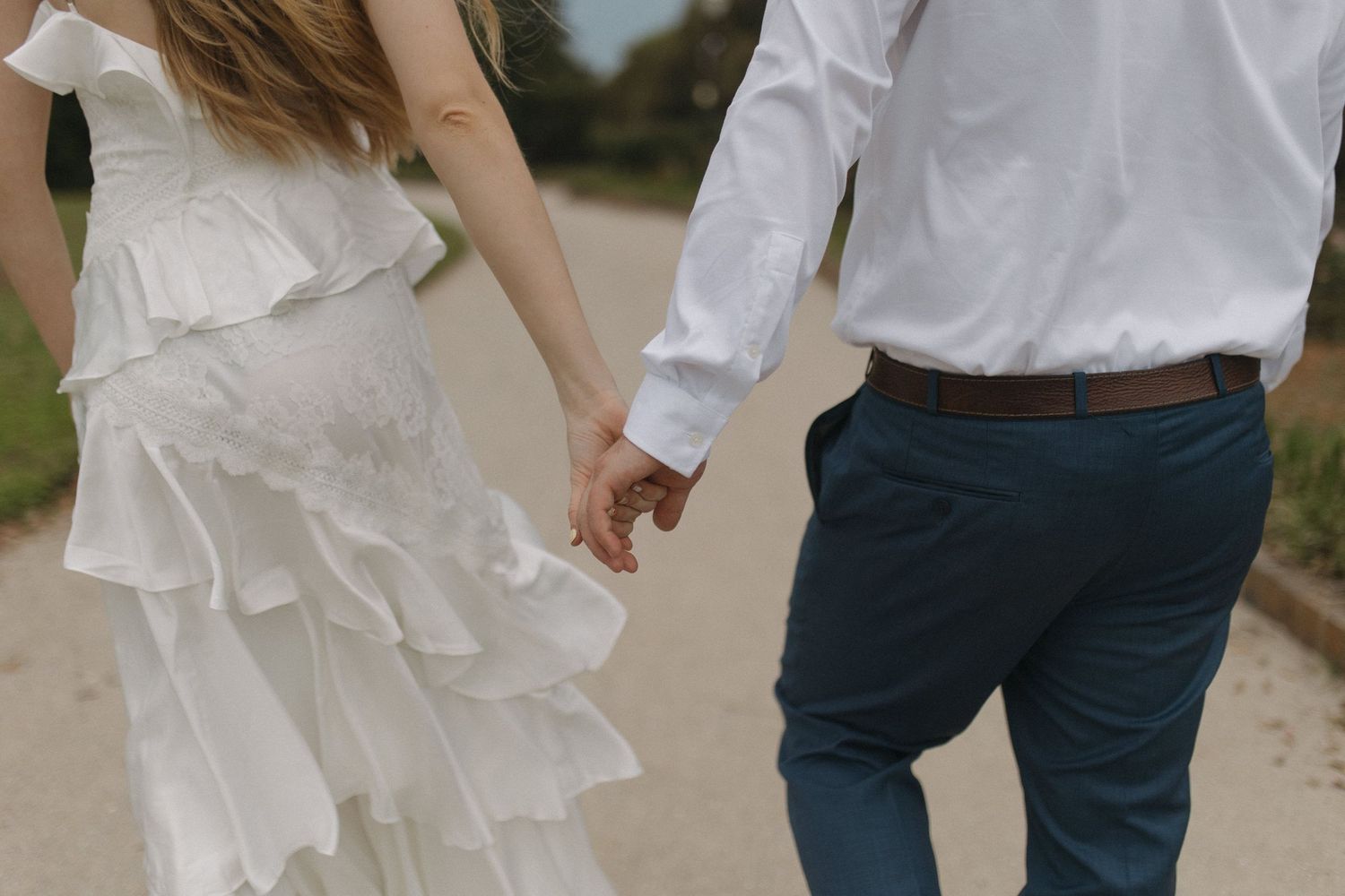 Bride in white ruffled dress and groom in blue pants holding hands while walking on a path.