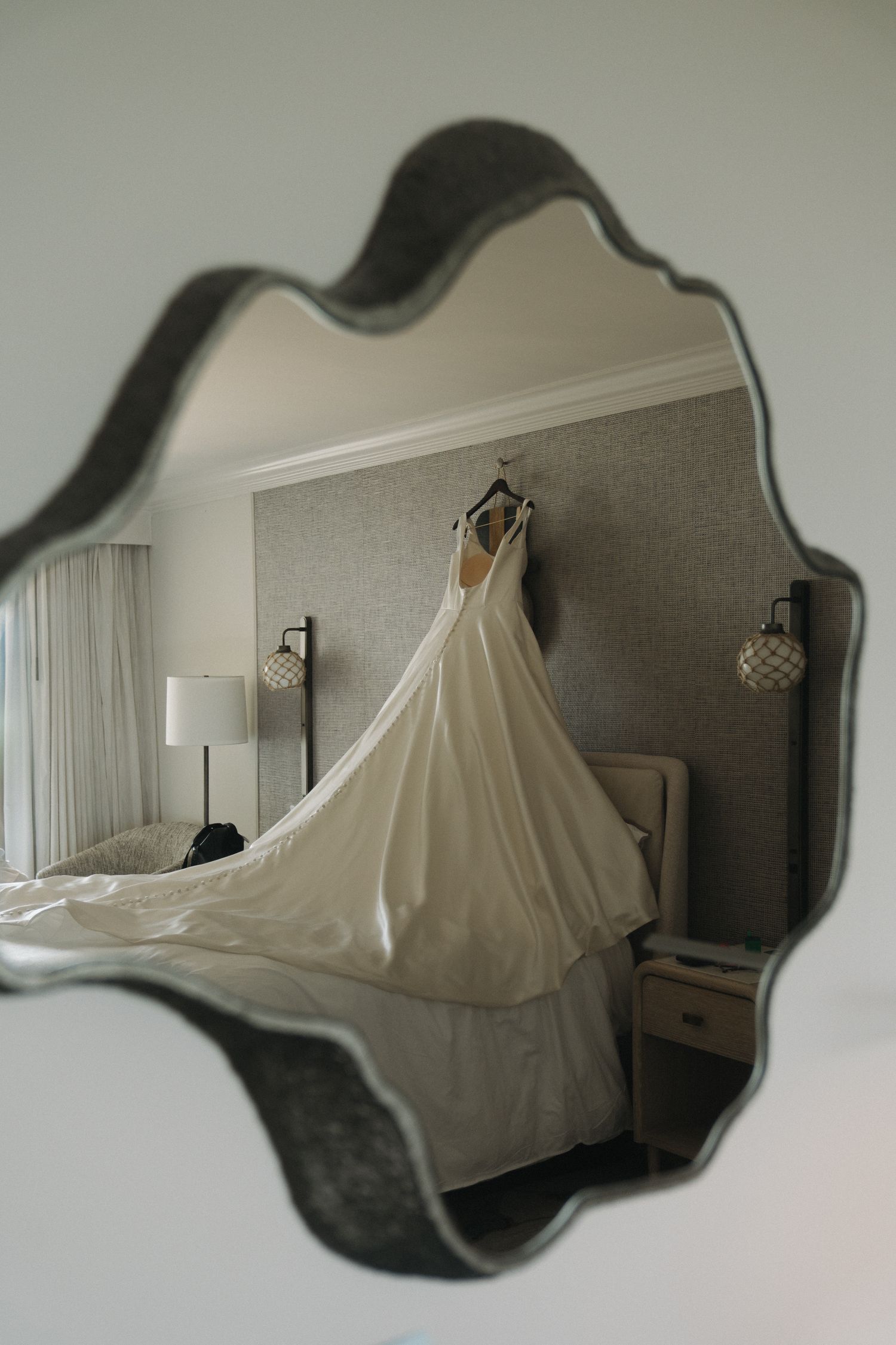 Elegant white wedding dress with a long train reflected in an ornate vintage mirror in a hotel room.