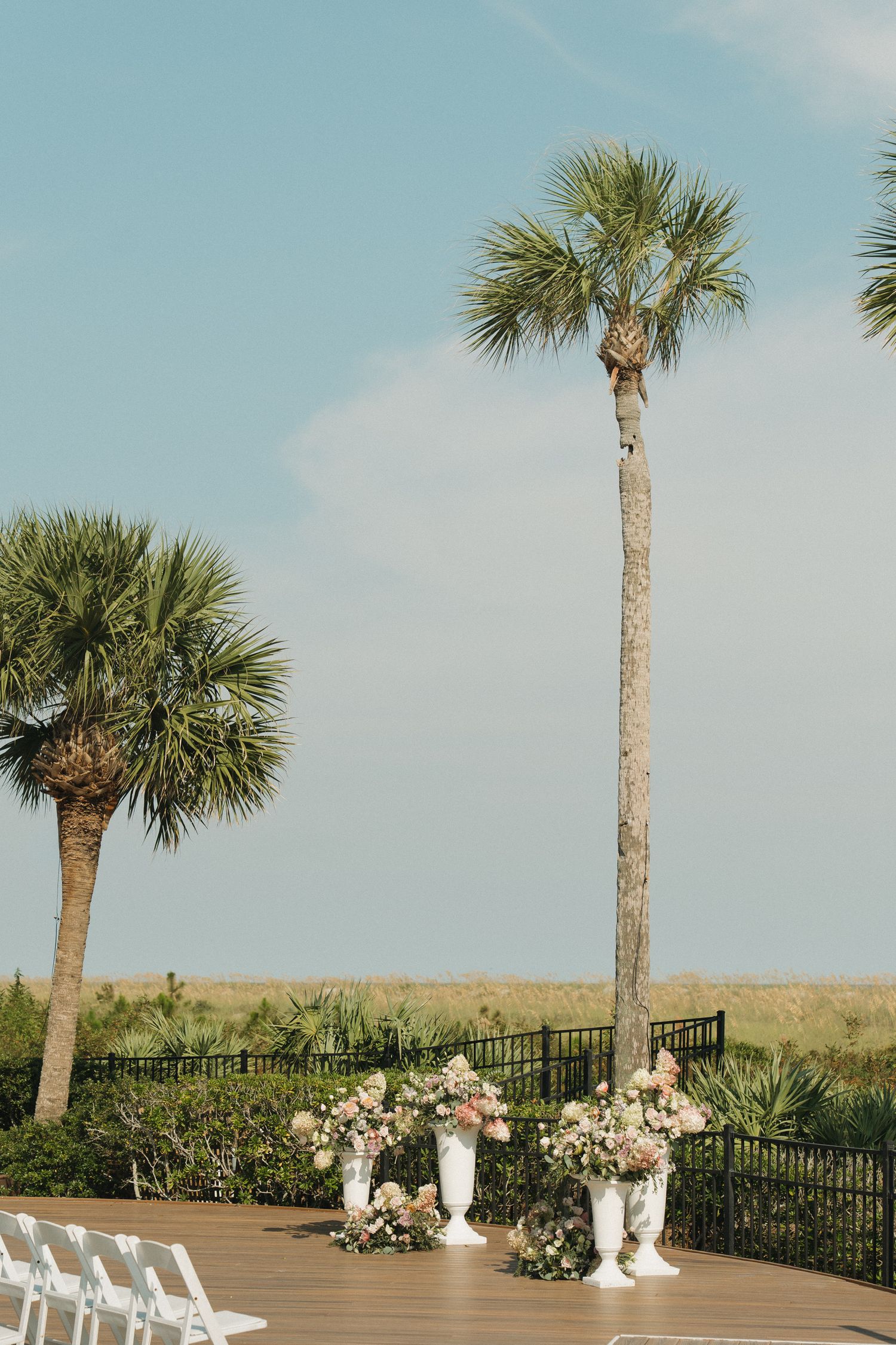 Outdoor wedding ceremony setup with white floral urns, palm trees, and marsh views under a blue sky.